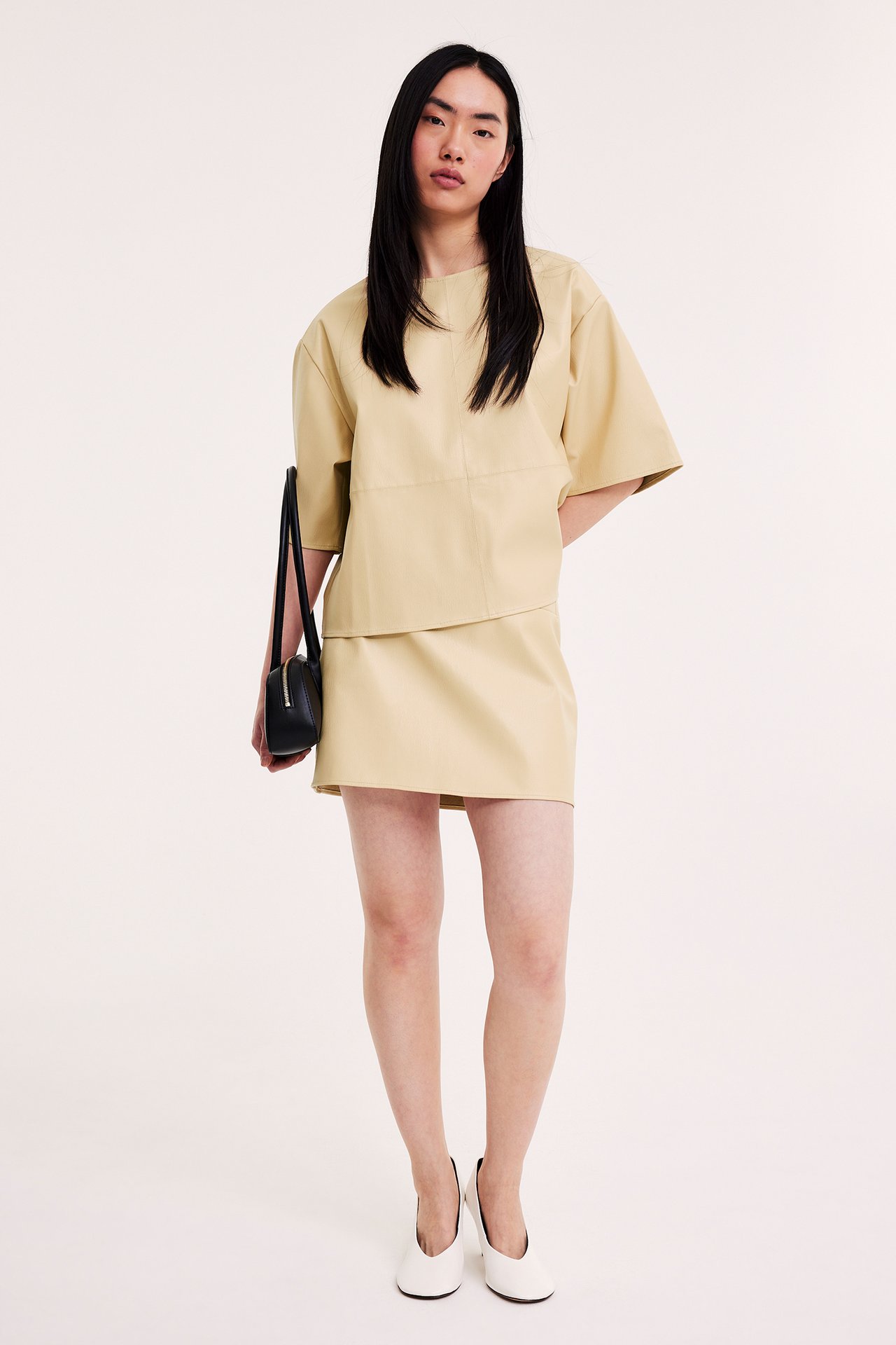 Edestä: Nainen vaaleankeltaisessa keinonahkahameessa ja topissa, mustassa laukussa, valkoisissa kengissä.