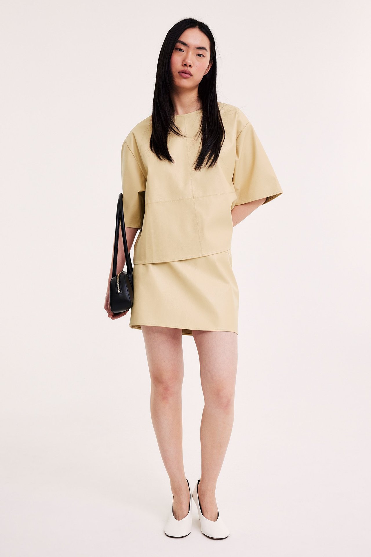 Edestä: Nainen vaaleankeltaisessa keinonahkahameessa ja topissa, mustassa laukussa, valkoisissa kengissä.