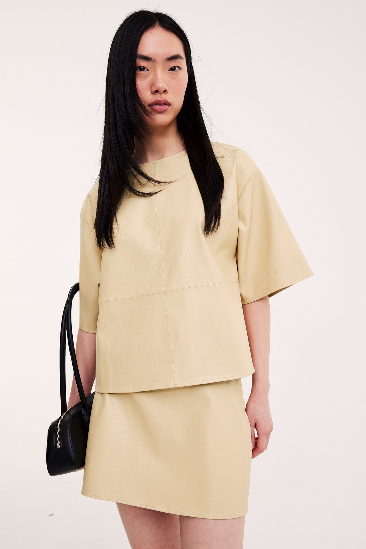 Etunäkymä naisesta, jolla on vaaleankeltainen lyhyt keinonahkahame, yhteensopiva toppi ja musta käsilaukku.