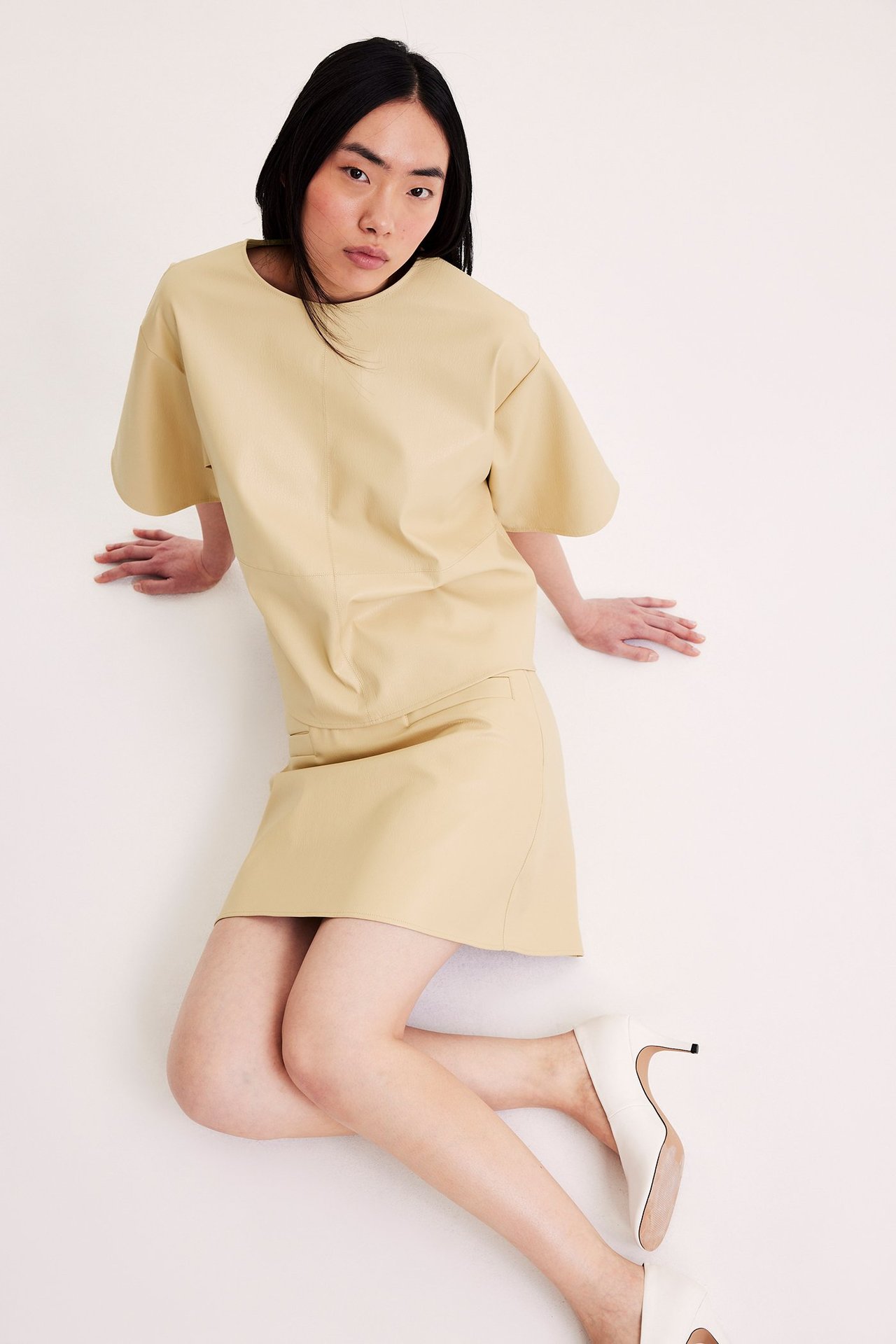 Edestä: Nainen vaaleankeltaisessa tekonahkaminihameessa, topissa ja valkoisissa koroissa.