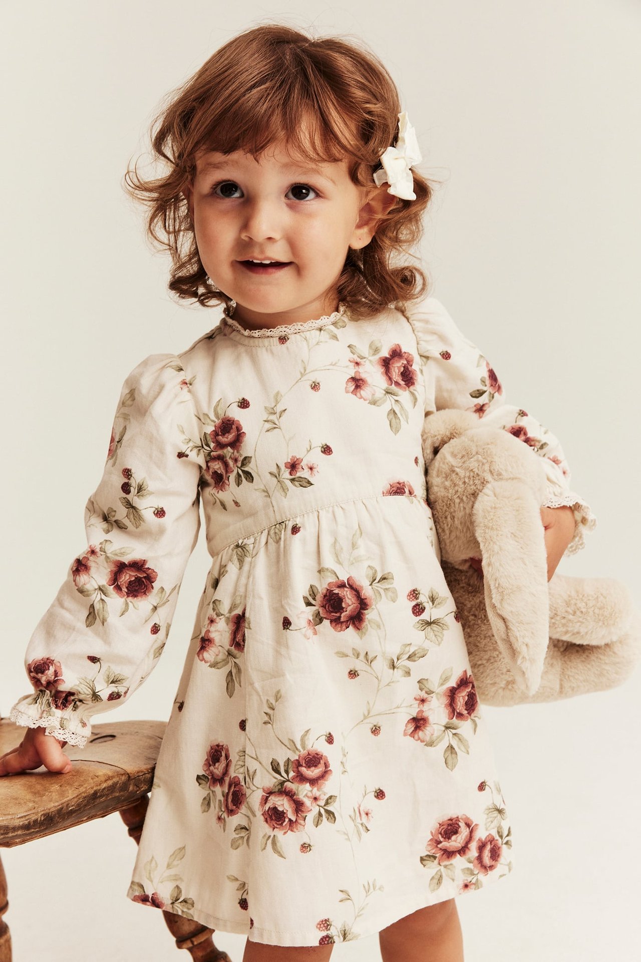 Kid bär en krämfärgad blommig långärmad klänning med spetsdetaljer, håller en beige plyschkanin.