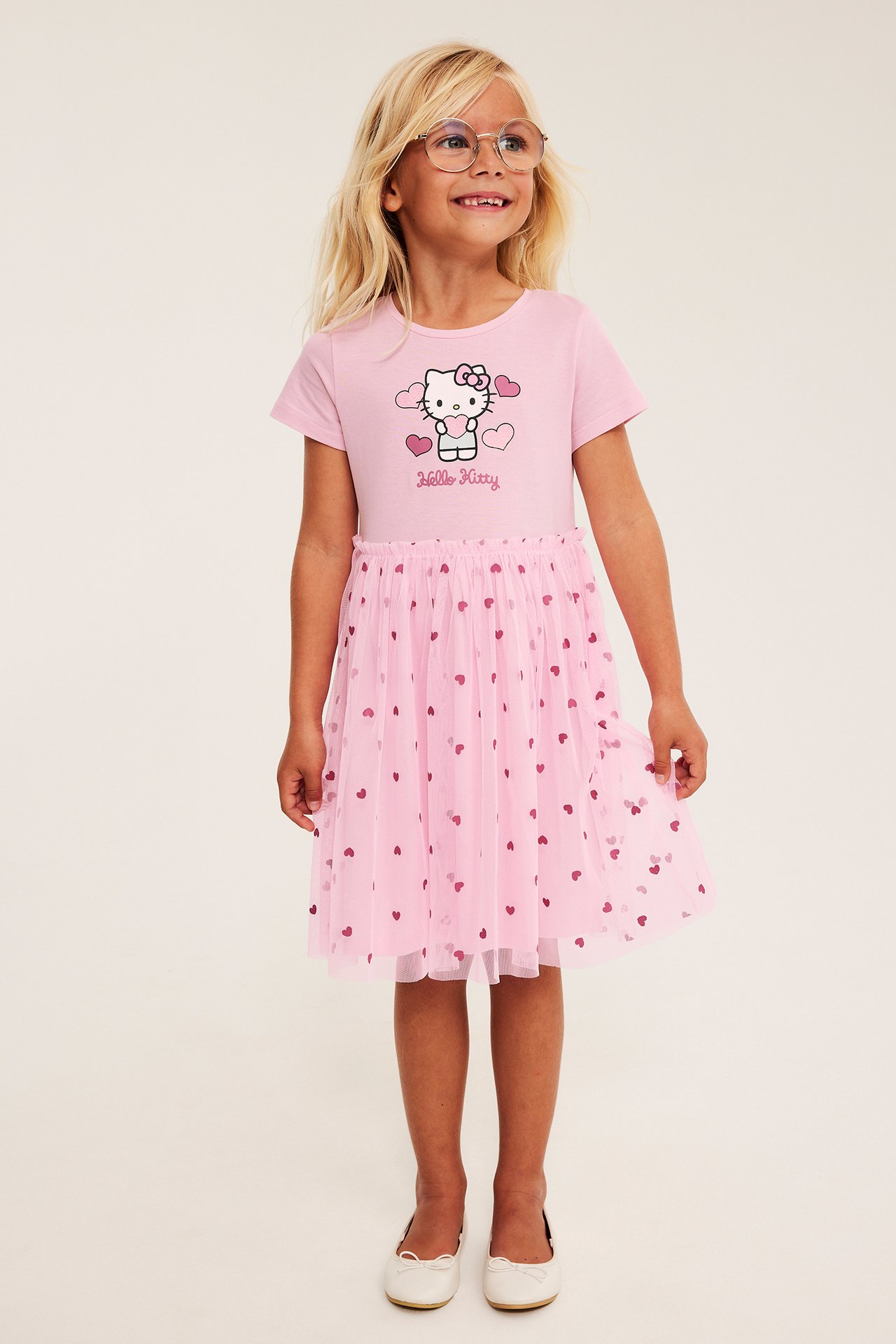 Barnrosa Hello Kitty-klänning med hjärttyllekjol, runda glasögon, vita ballerinaskor.