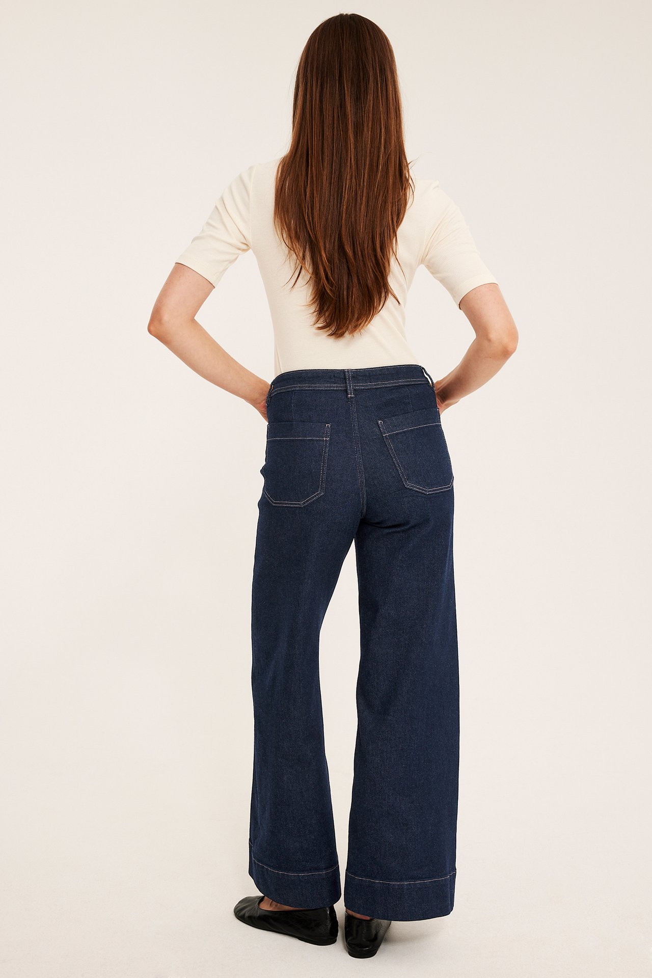 Ryggvisning av kvinne i mørkeblå vide jeans, offwhite kortermet topp og svarte flate sko.