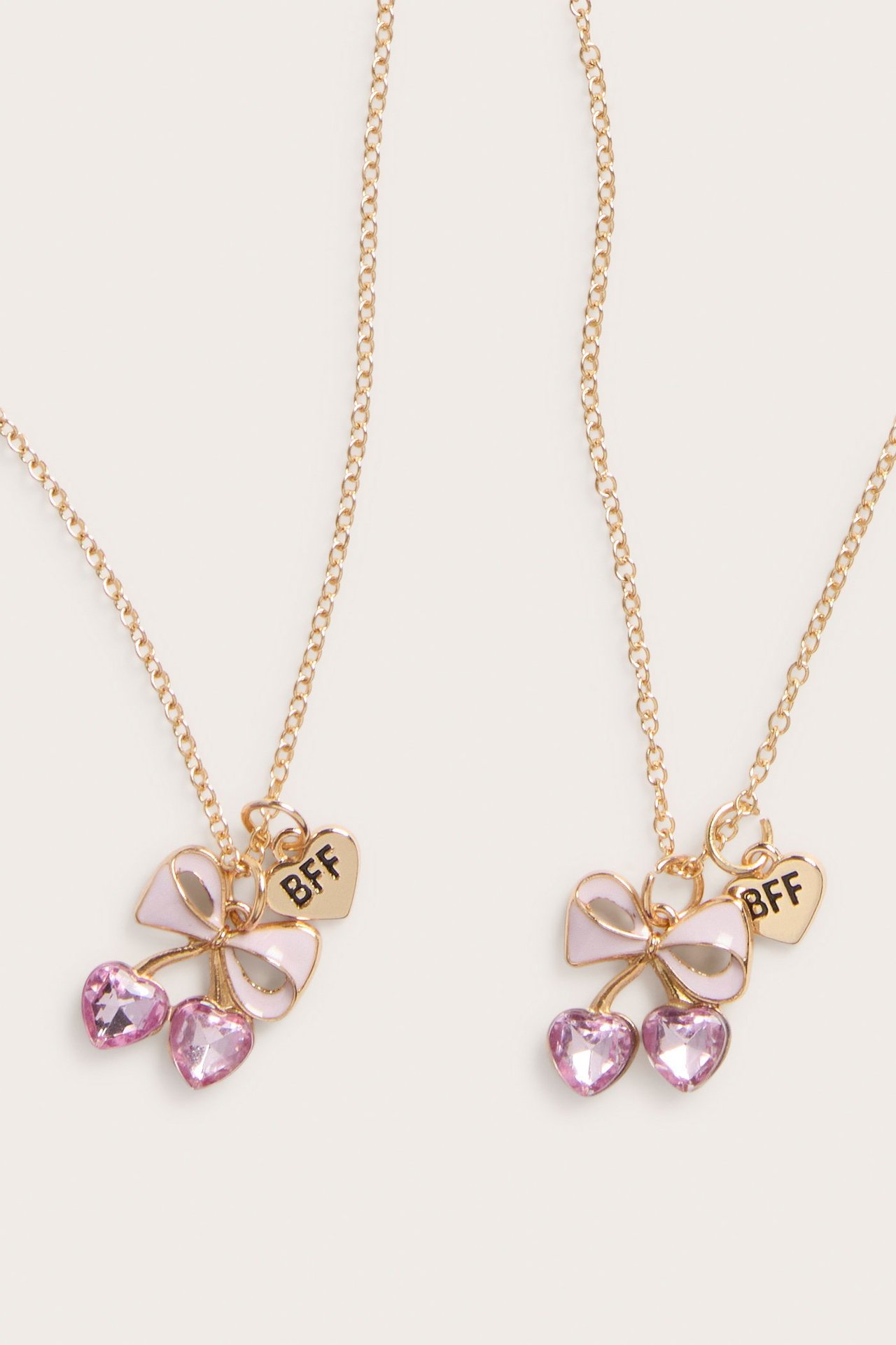 Nærbilde forfra av to gullfargede «BFF»-halskjeder med rosa sløyfer og hjerteformede krystaller.