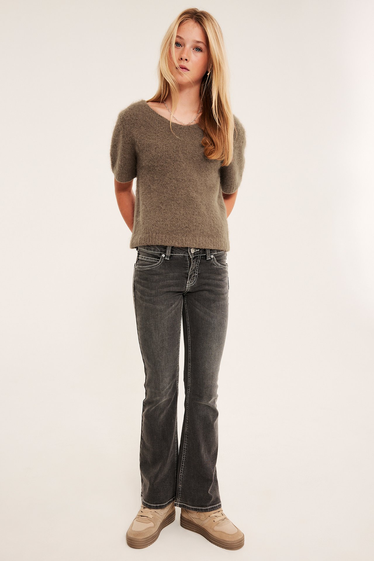 Frontbilde av en jente i en olivenfarget kortermet strikkegenser, mørkegrå flared jeans og beige sneakers.