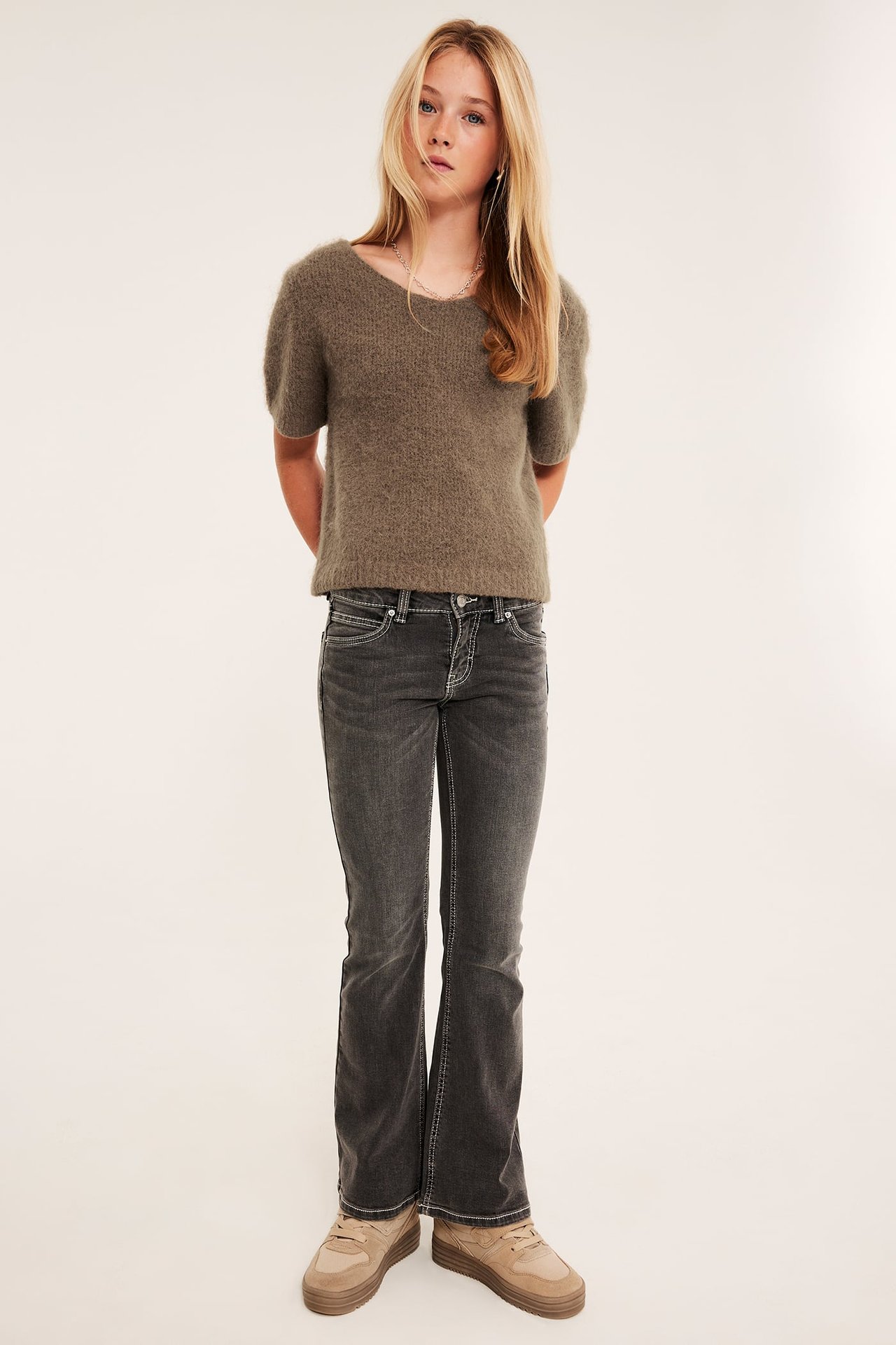 Frontbilde av en jente i en olivenfarget kortermet strikkegenser, mørkegrå flared jeans og beige sneakers.