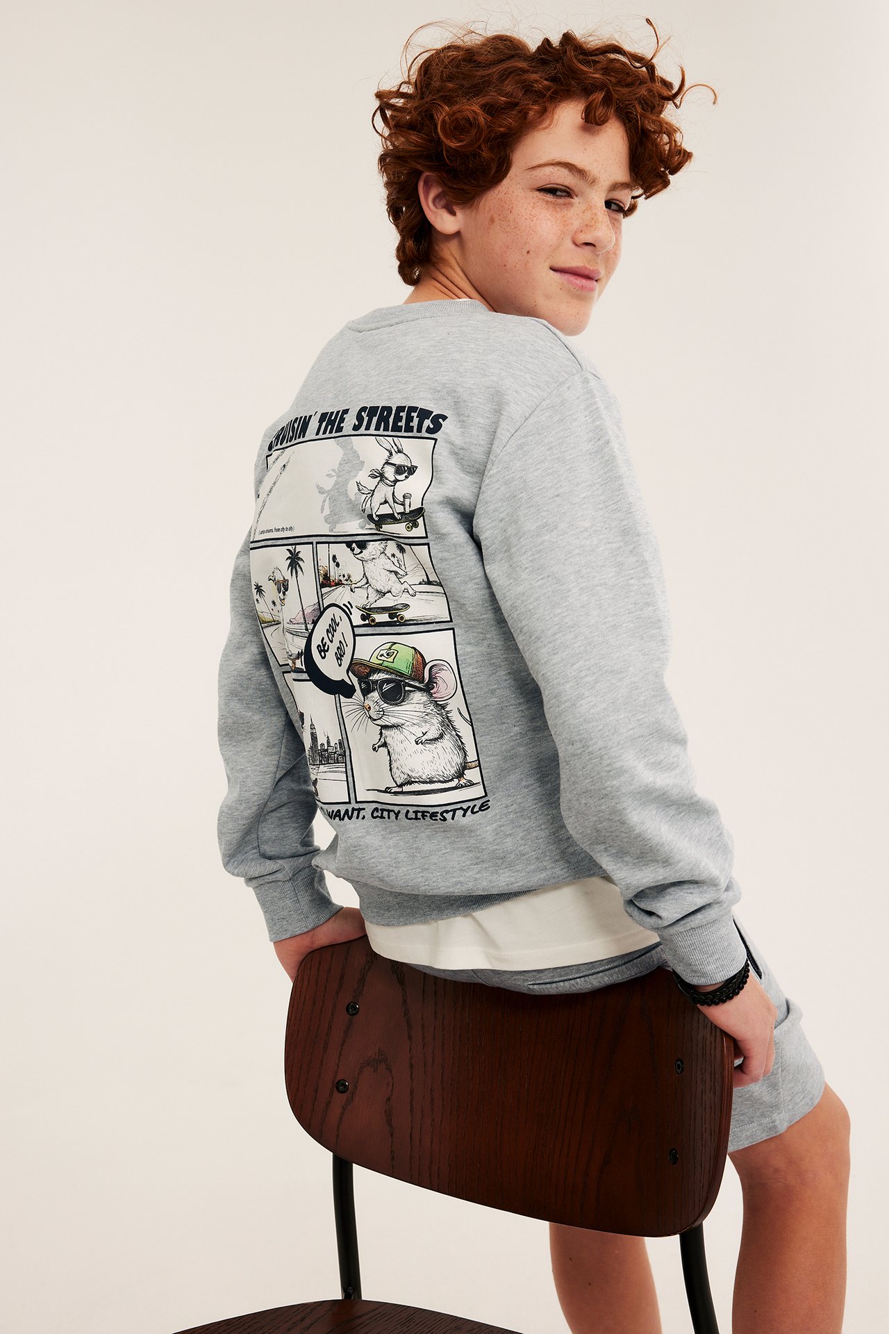 Bakfra: Barn i lys grå sweatshirt med tegneserie-dyretrykk, over hvit t-skjorte og grå shorts.