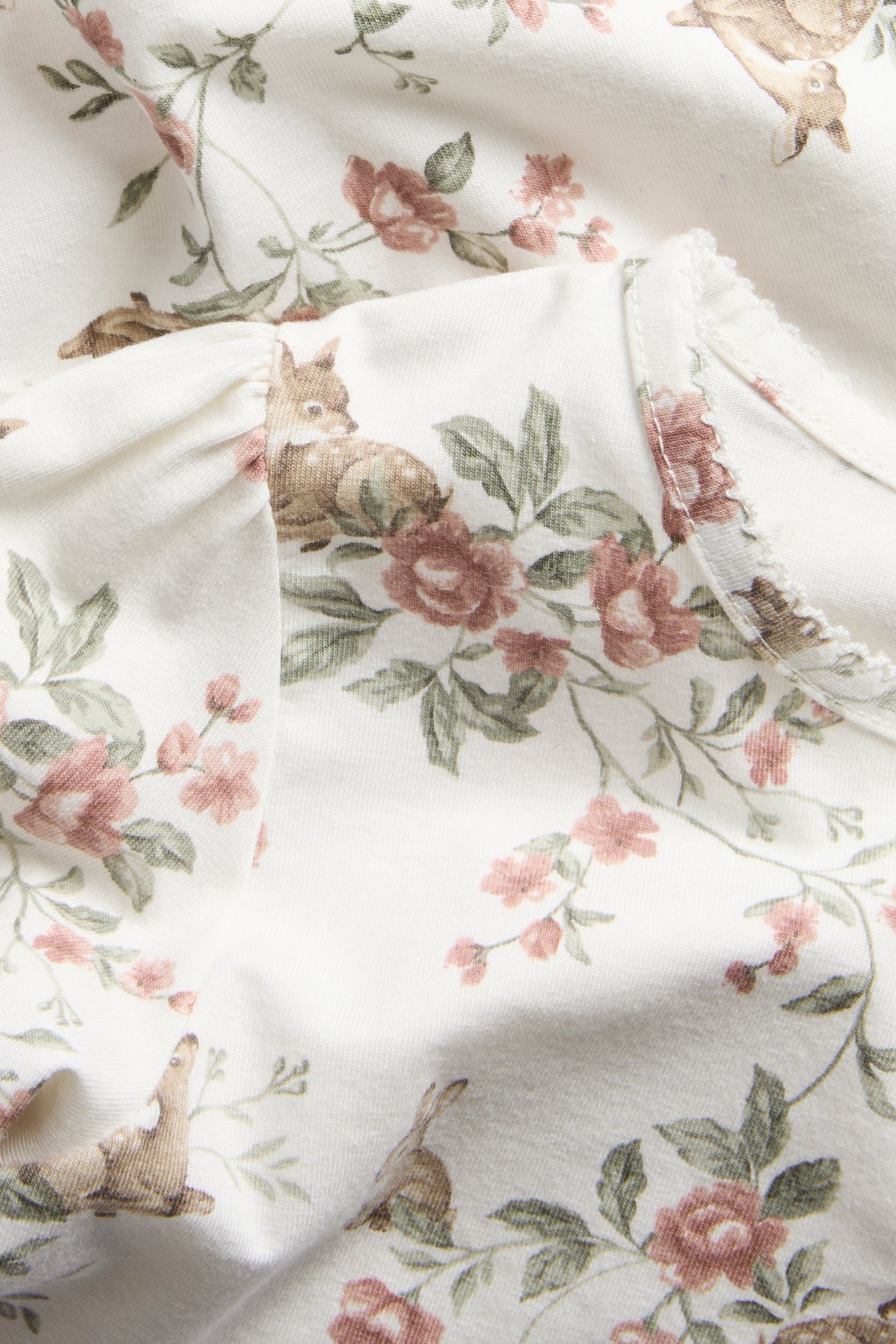 Nærbilde av offwhite langermet topp med rådyr- og blomstermønster, elastiske mansjetter og picotkant.