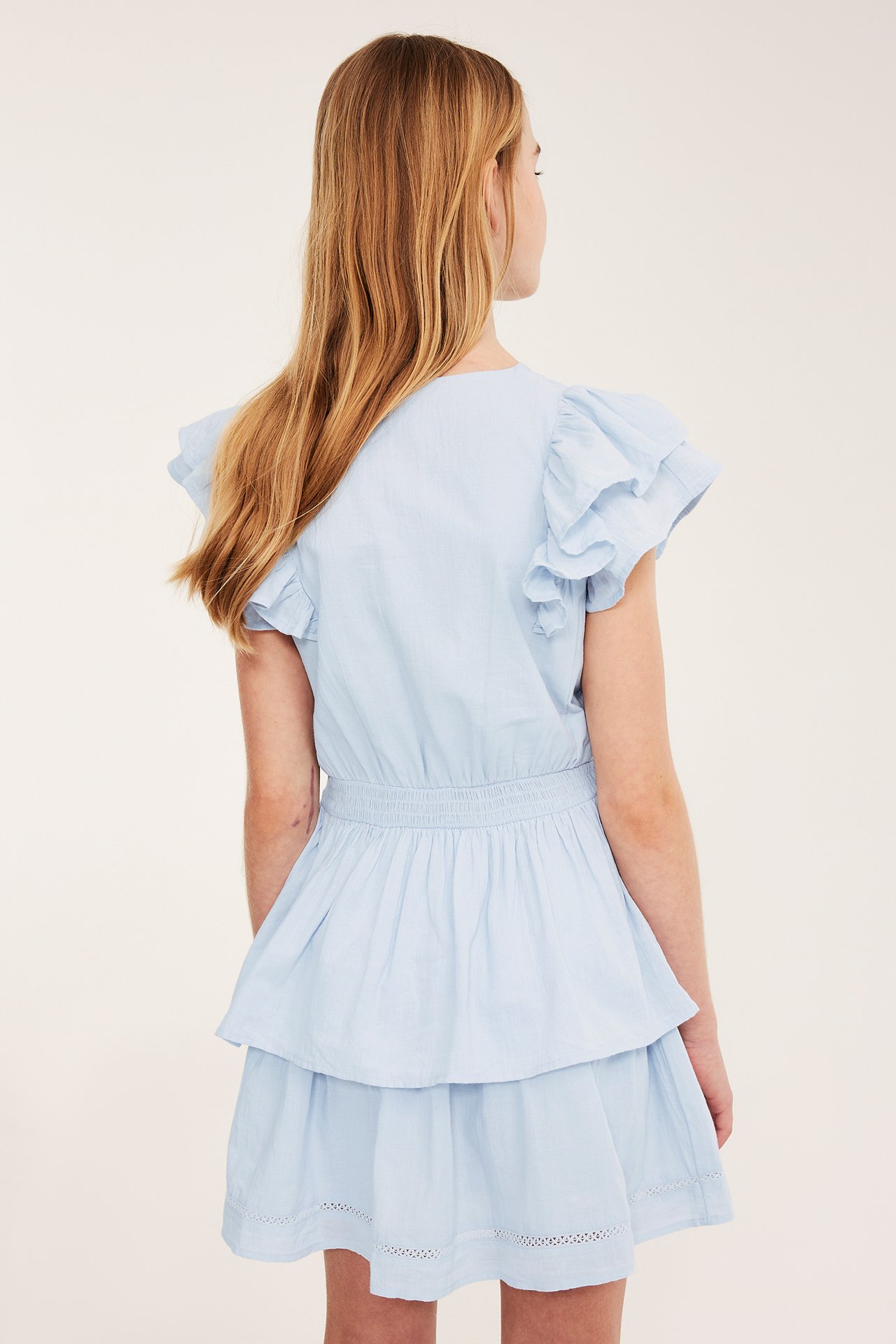 Widok z tyłu dziecka w jasnoniebieskiej bawełnianej sukience z falbanami, marszczoną talią i warstwową spódnicą.