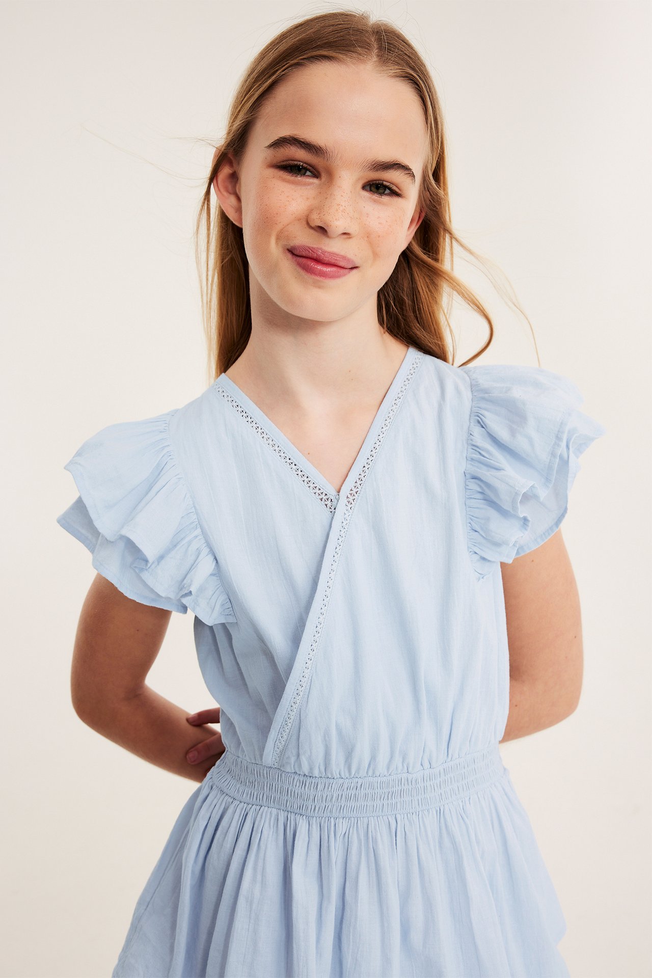 Widok z przodu: dziewczynka w jasnoniebieskiej bawełnianej sukience z falbanami, koronką i elastyczną talią.