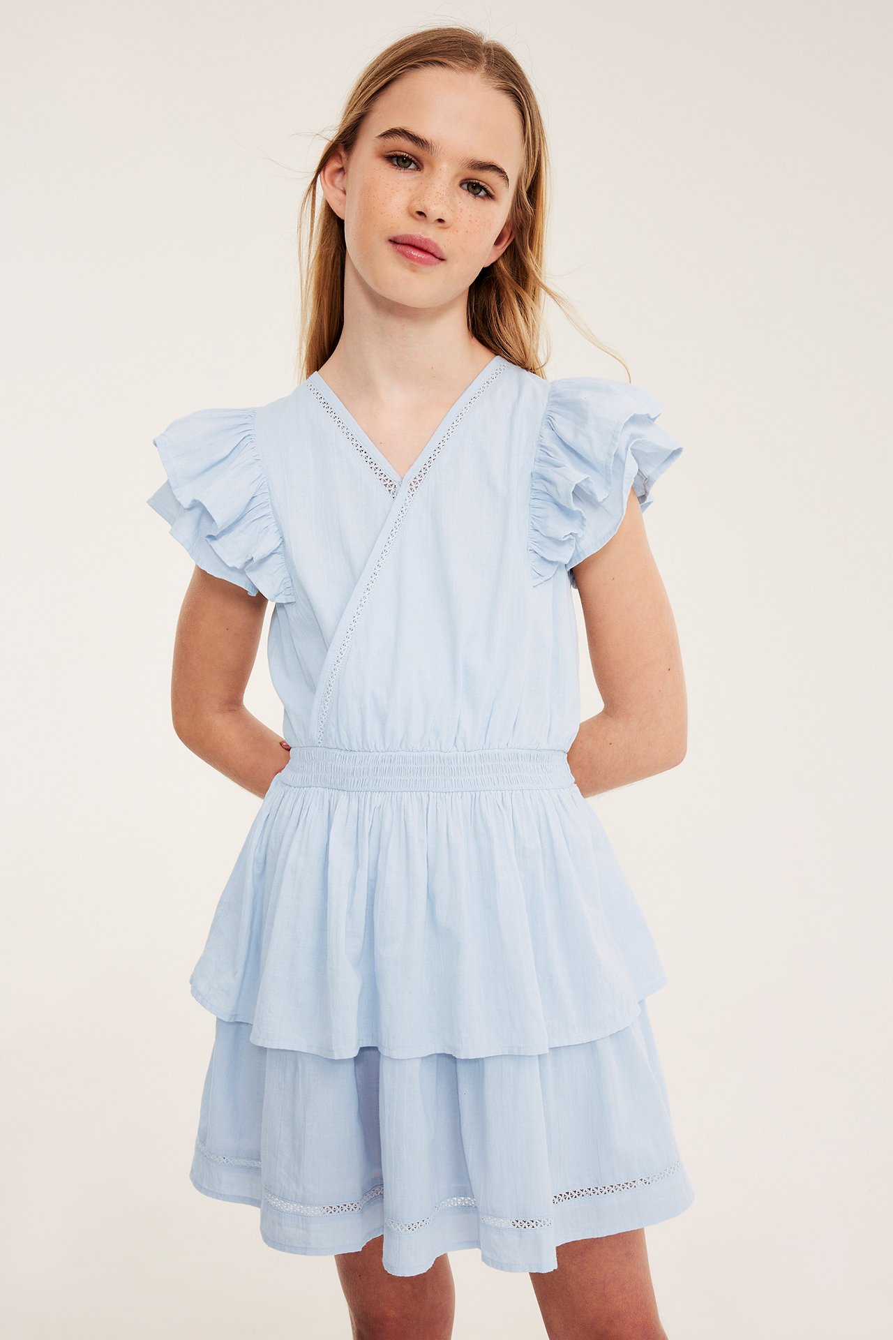 Edestäpäin kuvattu lapsi yllään vaaleansininen puuvillamekko, jossa röyhelöhihat, smokkirypytys ja kerrostettu helma.