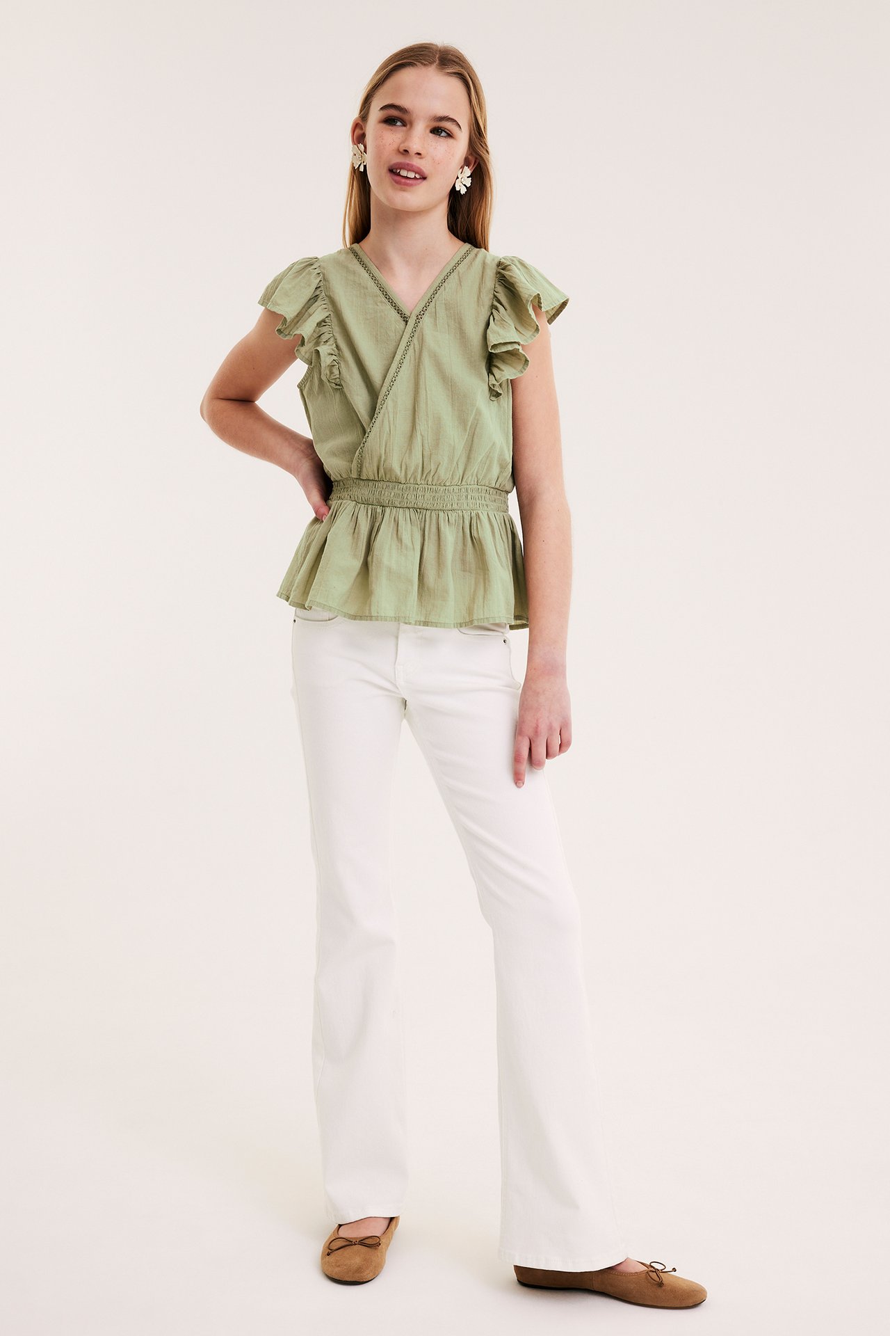 Frontvy av ett barn i mörkgrön bomulls volangblus med smockmidja, vita byxor och bruna ballerinaskor.