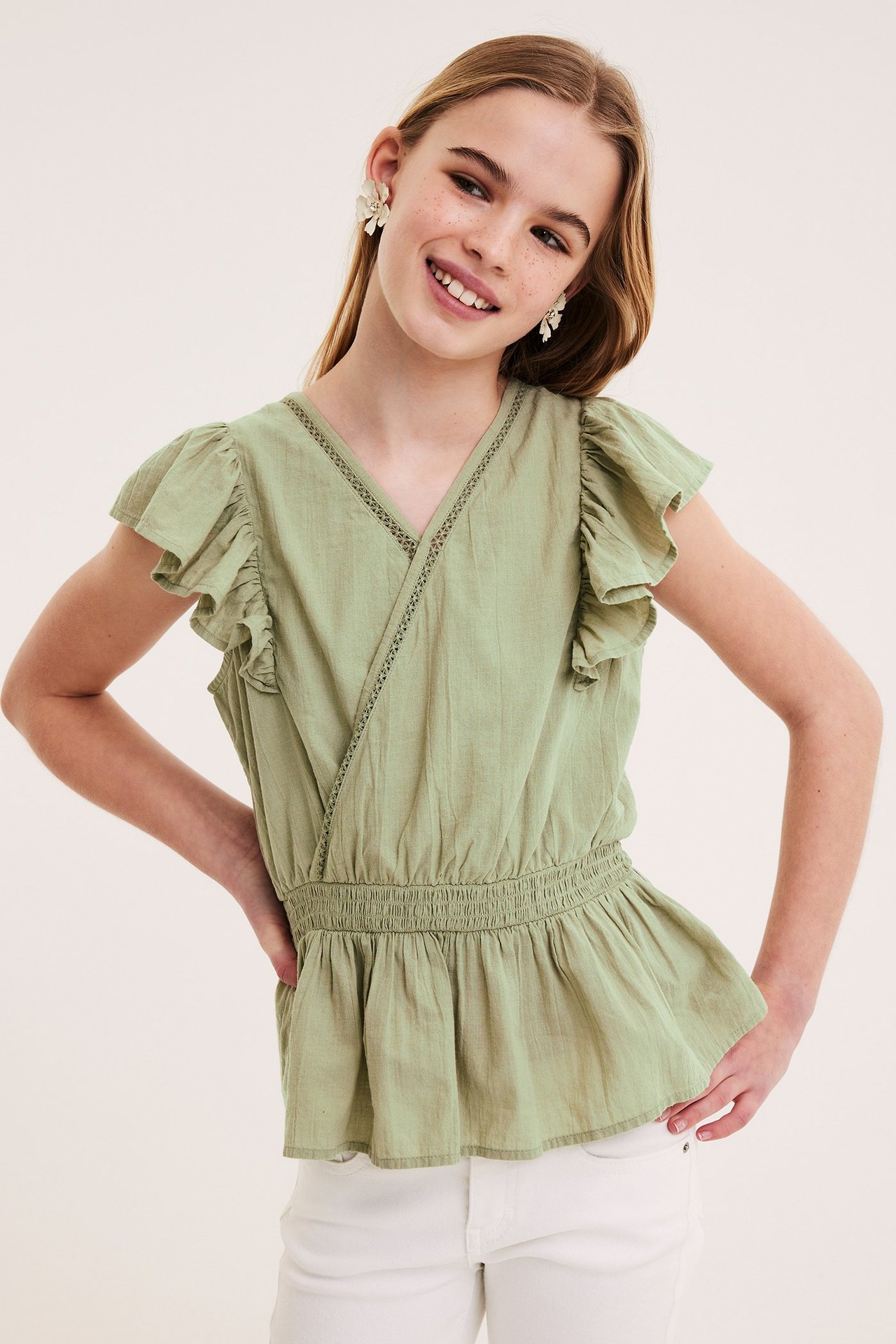 Barn bærer mørkegrønn bomullsbluse med volanger, smock i midjen, hvite bukser og blomsterøredobber.