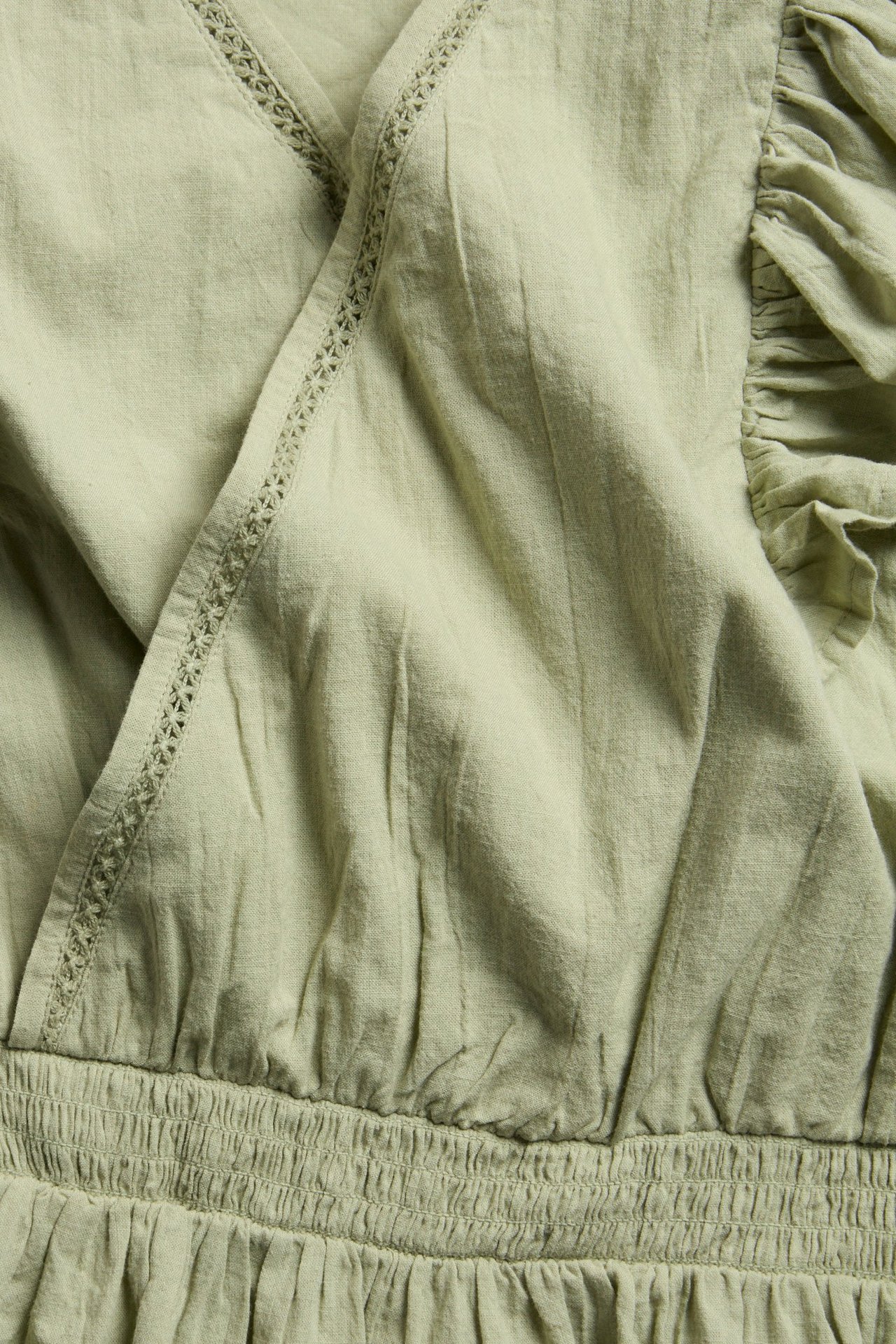 Närbild av mörkgrön bomullsblus med omlottring, spetsdetaljer, volanger och smock i midjan.