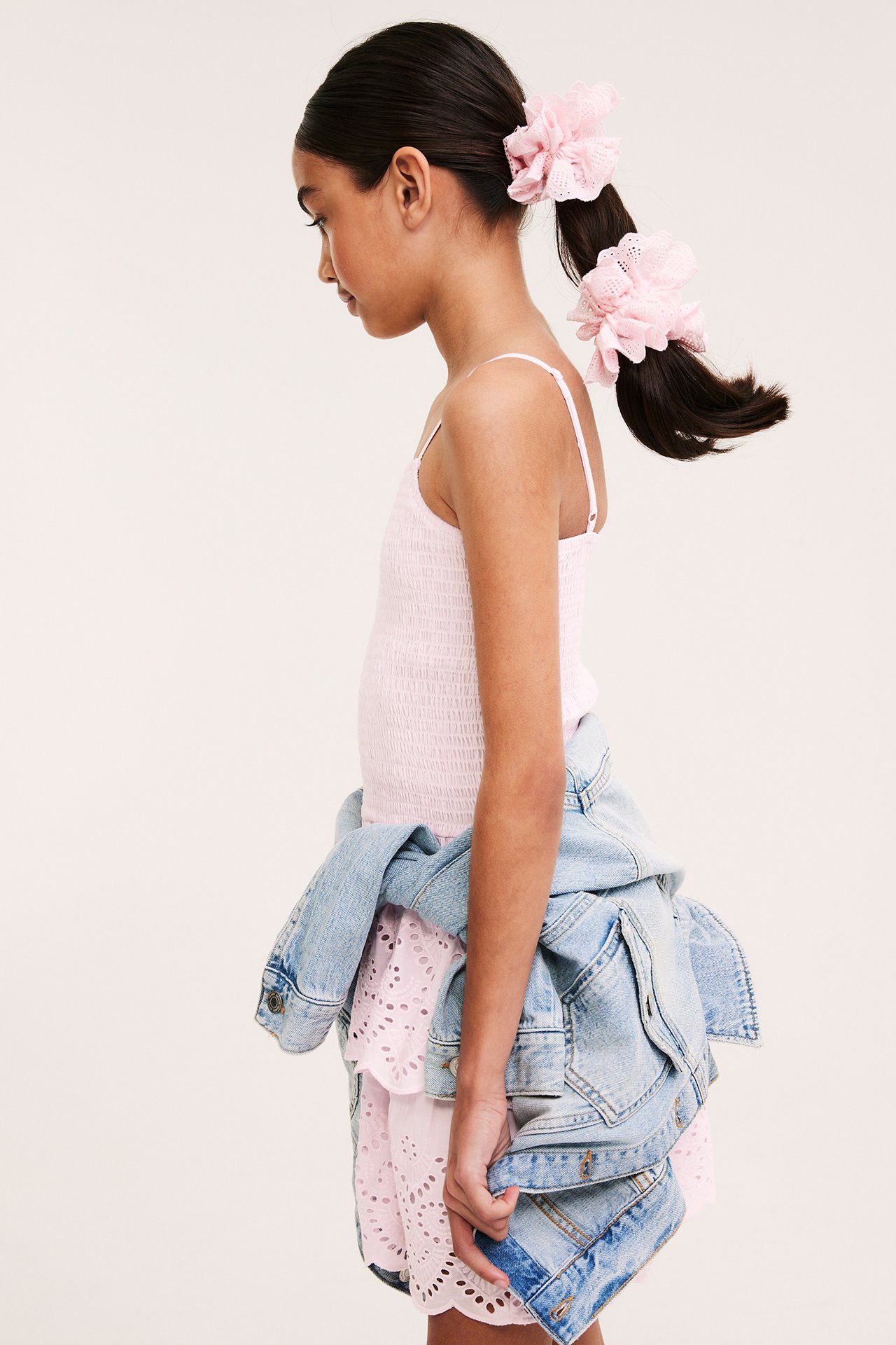 Sidovy: Barn i ljusrosa smockad broderie anglaise-klänning, jeansjacka, rosa hårband.