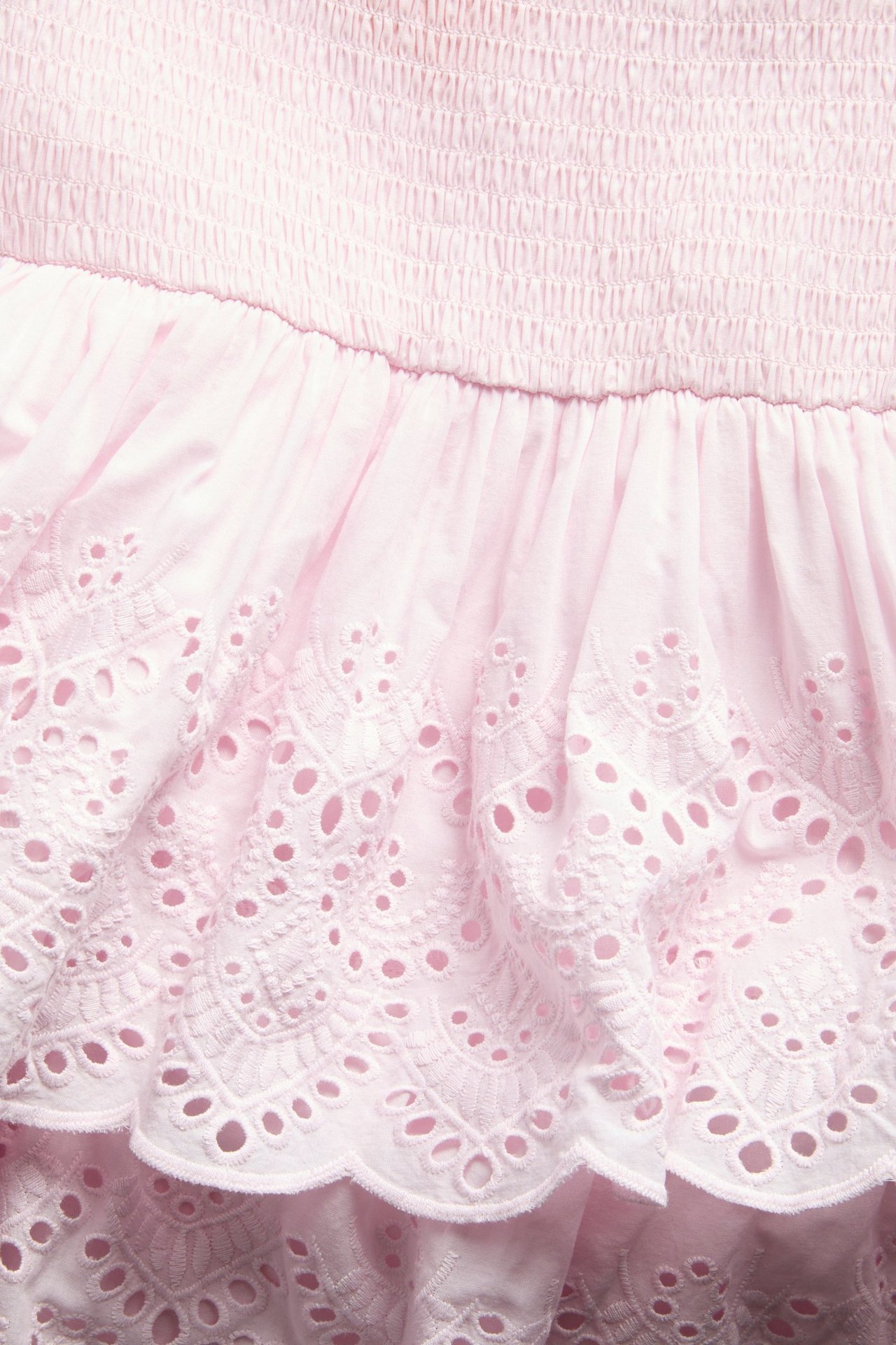 Närbild av en ljusrosa bomullsklänning med smockat liv och volanger med broderie anglaise.