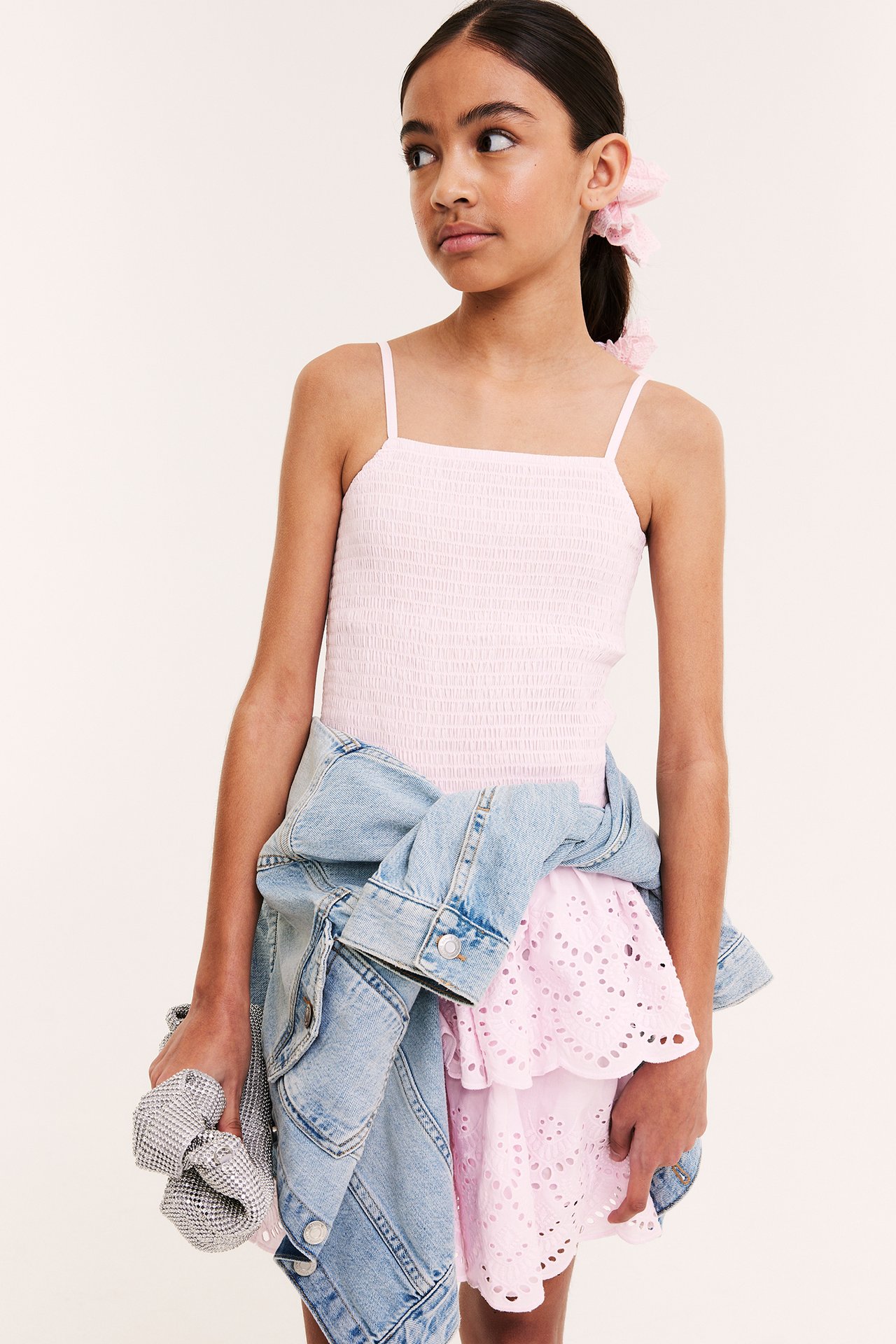 Framifrån: tjej i ljusrosa smockad bomullsklänning med broderie anglaise, håller jeansjacka och silverscrunchie.