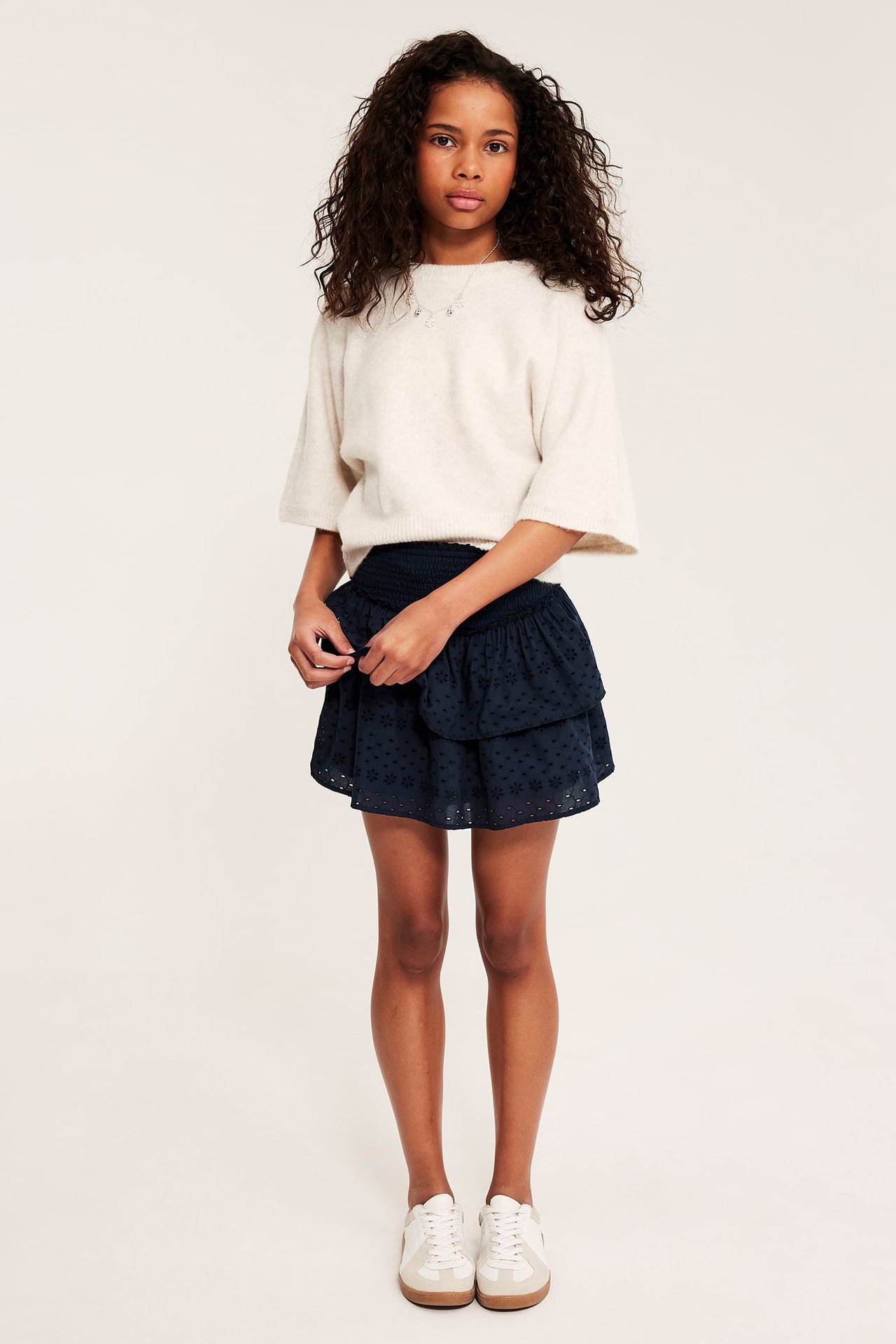 Frontutsikt av barn i marineblått broderie anglaise-skjørt med smockmidje, off-white genser og joggesko.