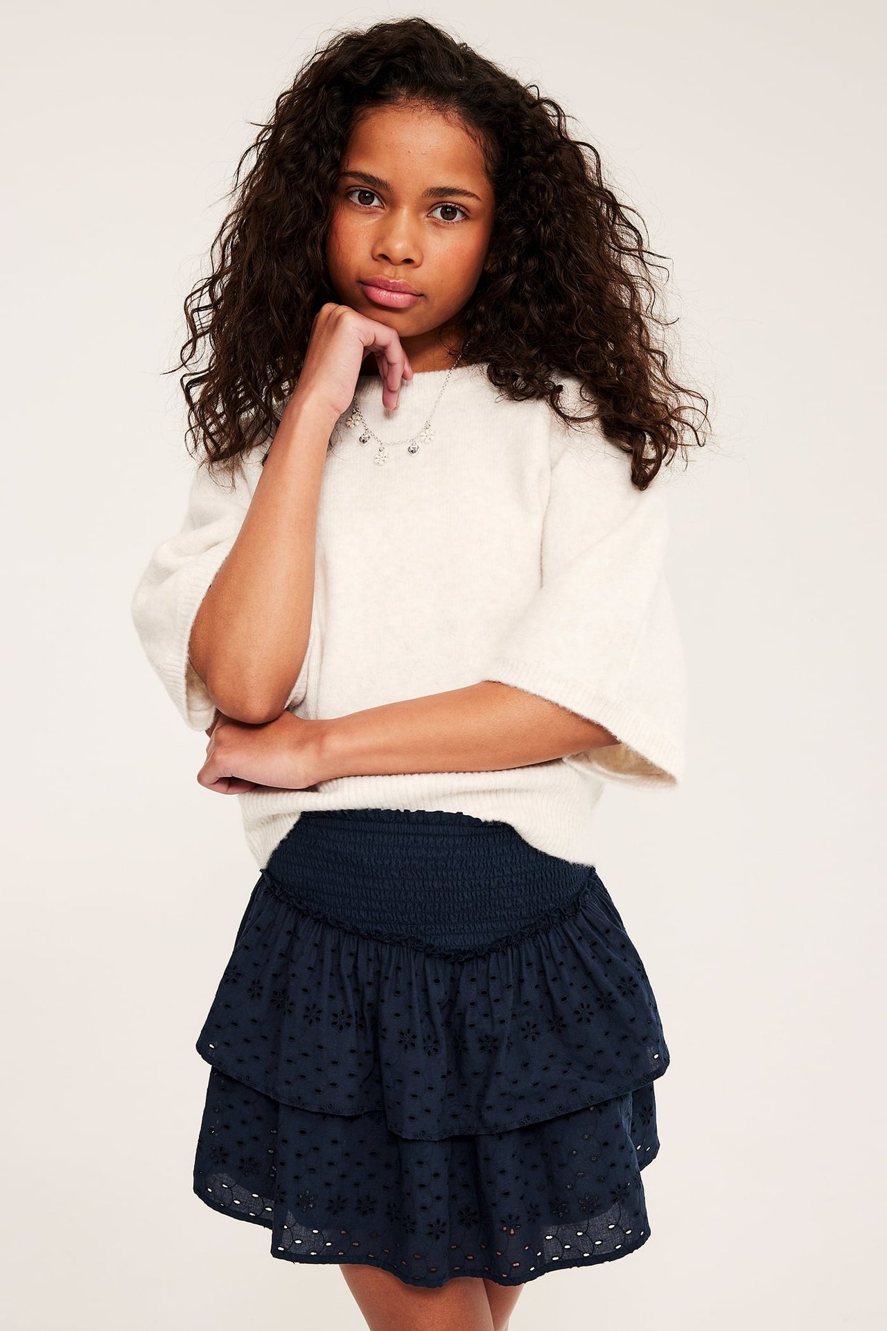 Widok z przodu dziecka w ciemnoniebieskiej spódnicy z haftem angielskim, marszczonej w pasie i jasnobeżowym swetrze.