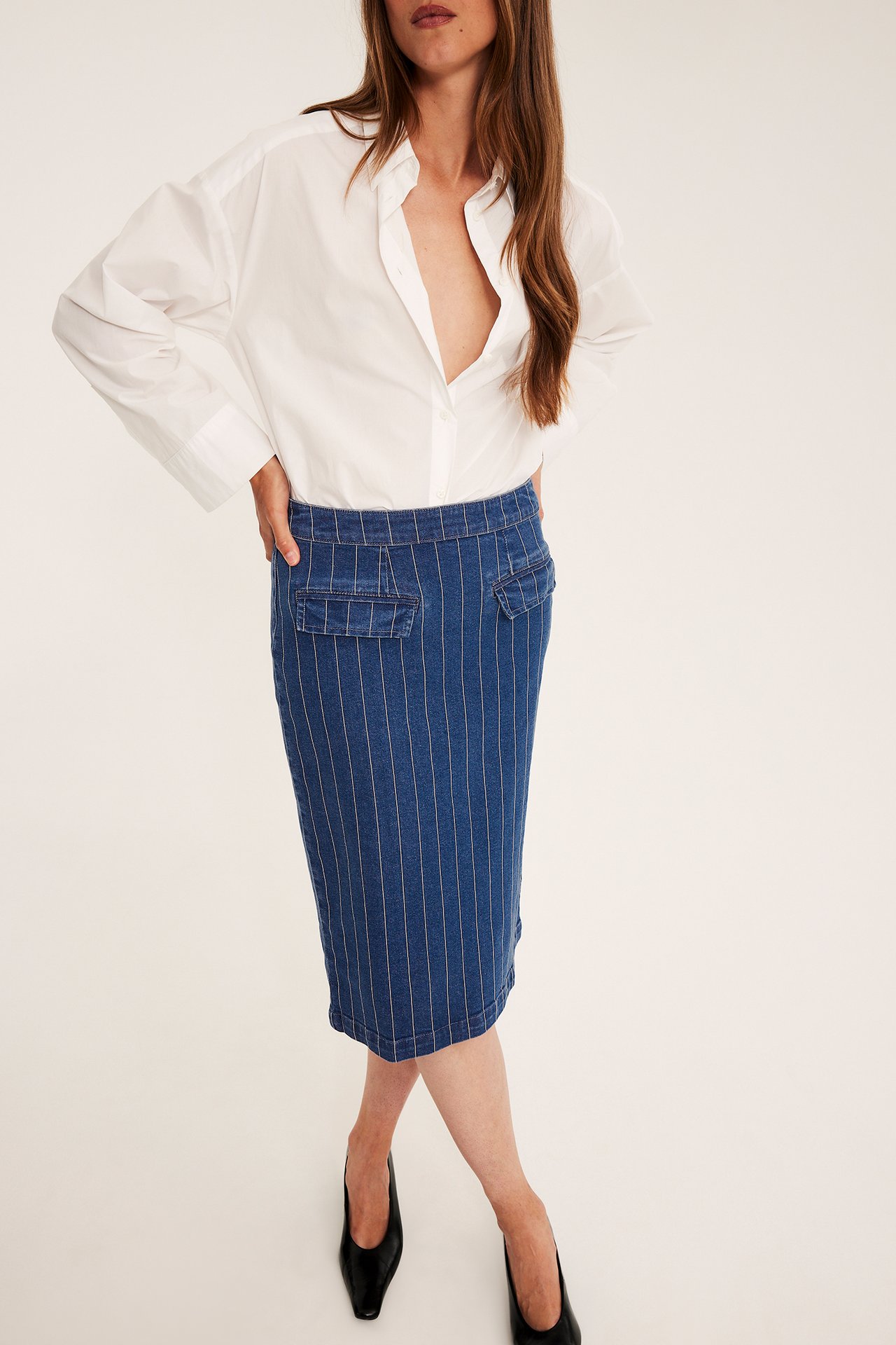 Forfra, kvinne iført blå pinstripe denim blyantskjørt med lommer, hvit skjorte og svarte hæler.