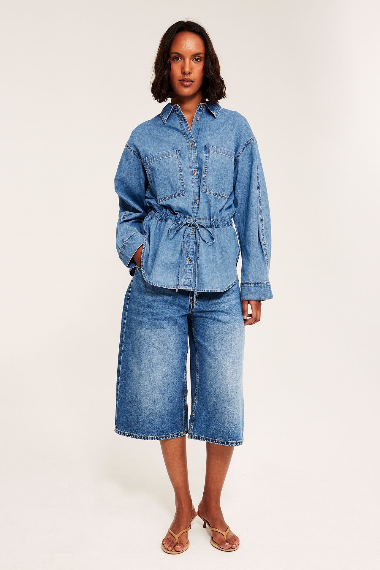 Framifrån, kvinnlig modell i blå jeansskjorta med dragsko, matchande culottes och sandaler.