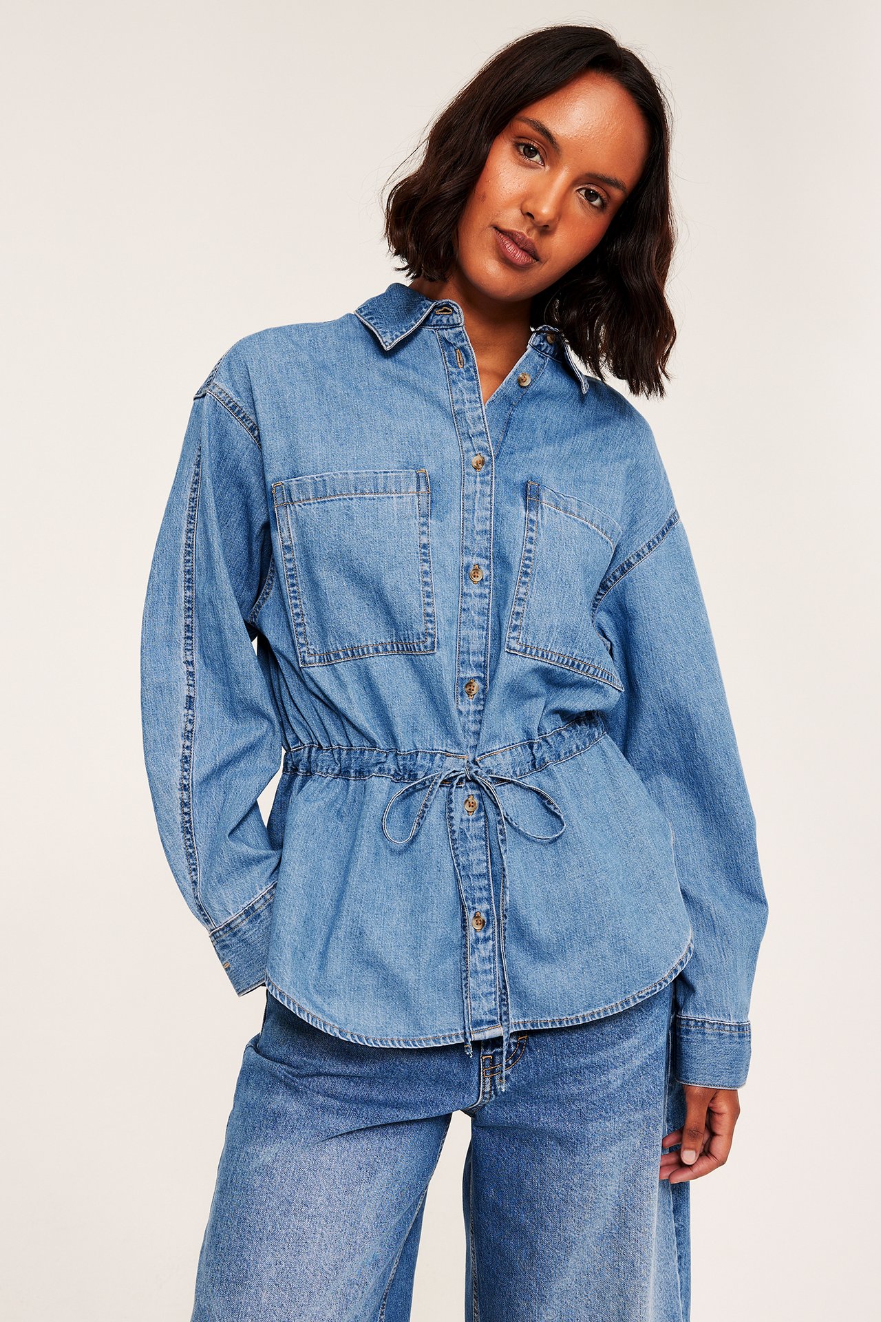 Framifrån, kvinna i blå denimskjorta med dragsko i midjan över blåa jeans.
