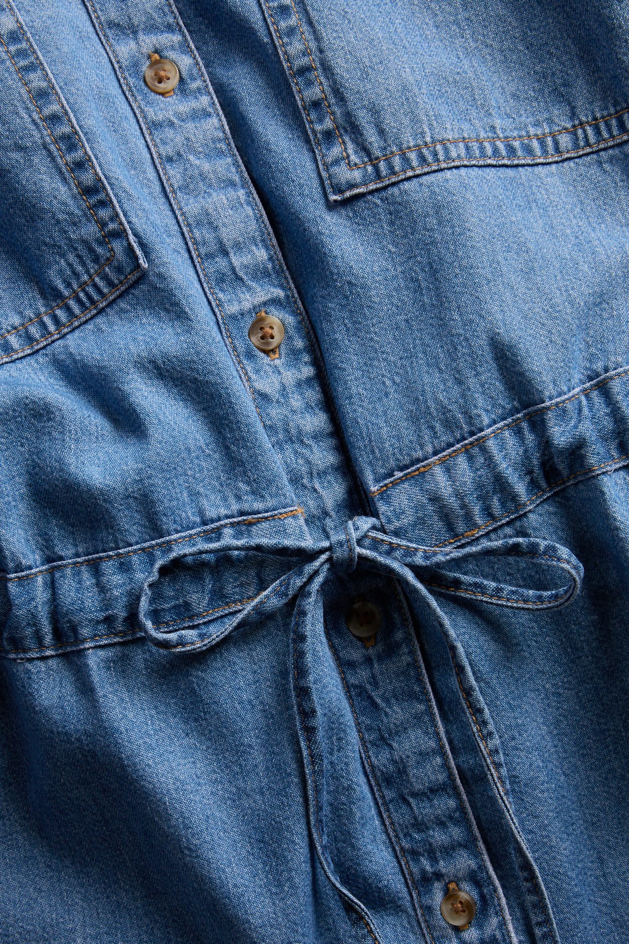 Närbild på blå denimskjortklänning med knappar fram och knytband i midjan.