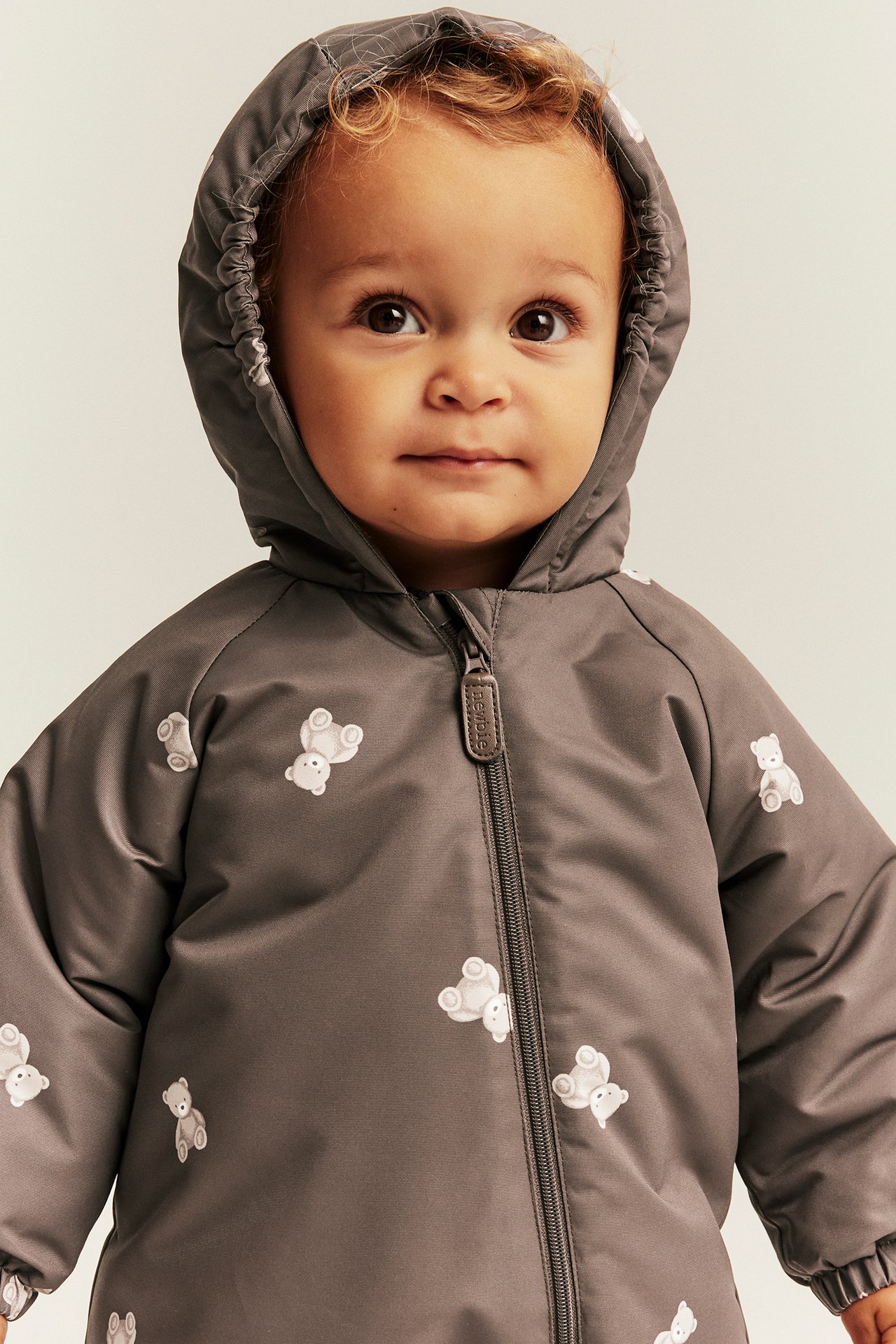 Nærbilde av baby i grå hetteoverall med hvitt teddybjørntrykk, glidelås foran.