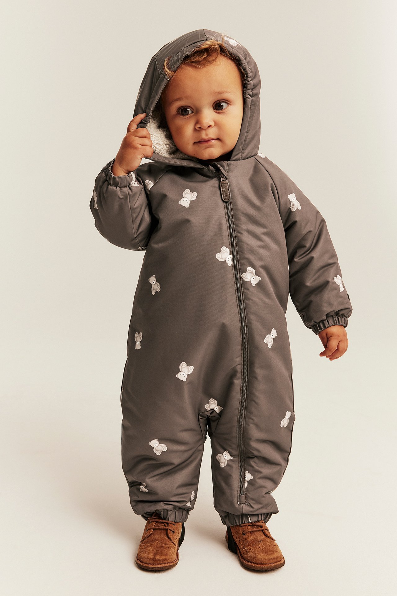 Frontbilde av baby i mørk grå hetteoverall med hvite bamser og brune sko.