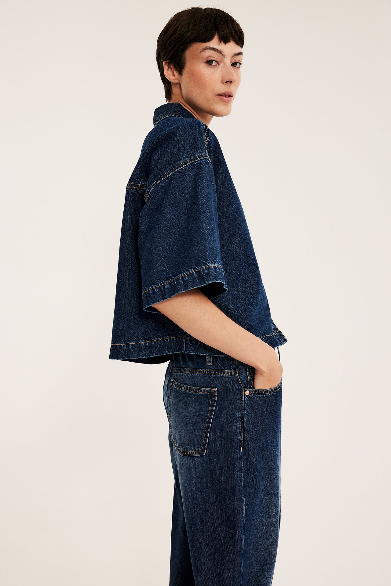 Naispuolinen malli sivukuvassa pukeutuneena tummansiniseen lyhythihaiseen denim-crop-paitaan ja leveälahkeisiin housuihin.