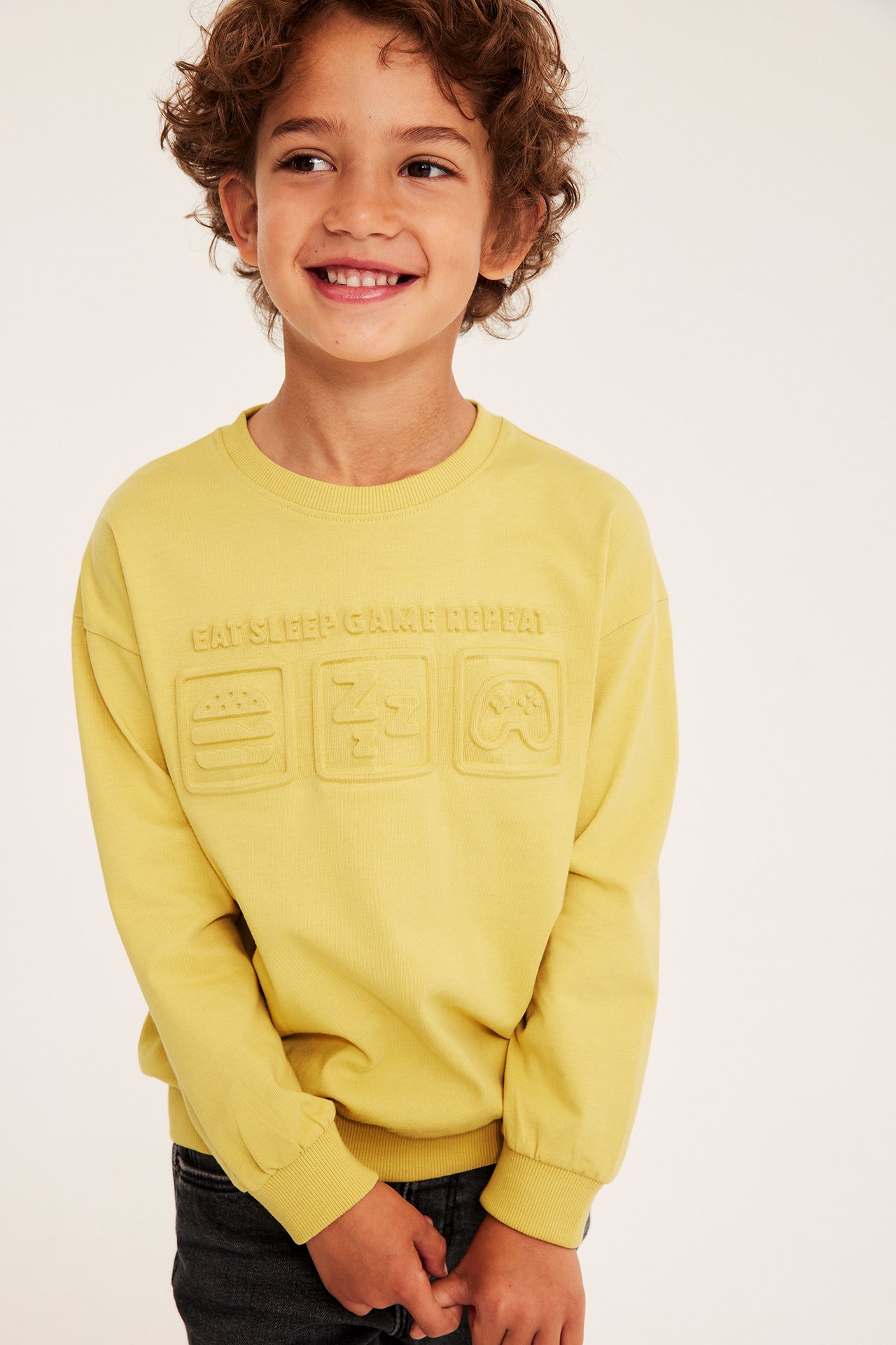 Framifrån, ett leende barn i en gul sweatshirt i bomull med 3D-effekttryck, samt svarta jeans.