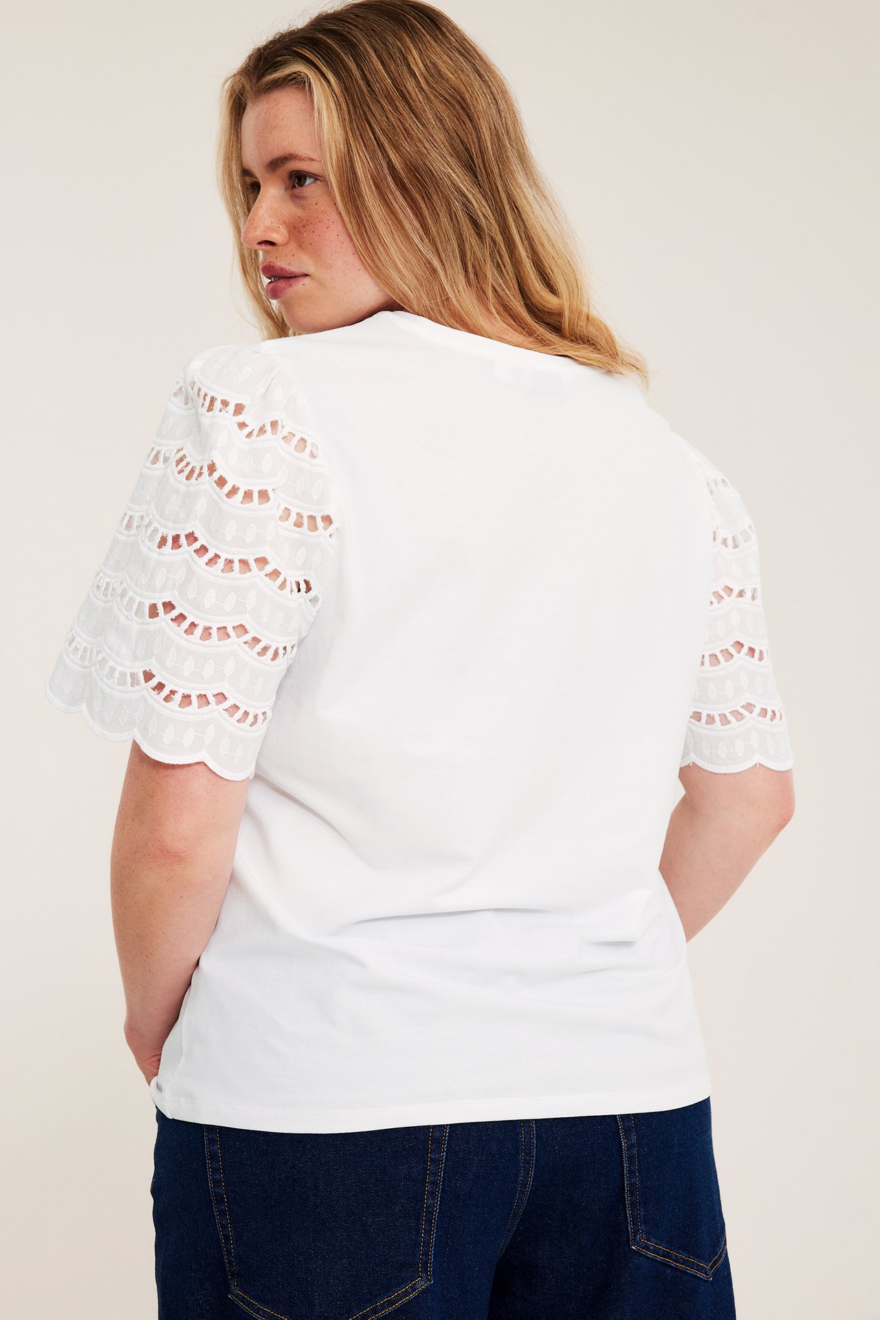 Naisen takakuva valkoisessa lantiopituisessa topissa, jossa on puuvillaiset broderie anglaise -hihat ja tummat farkut.