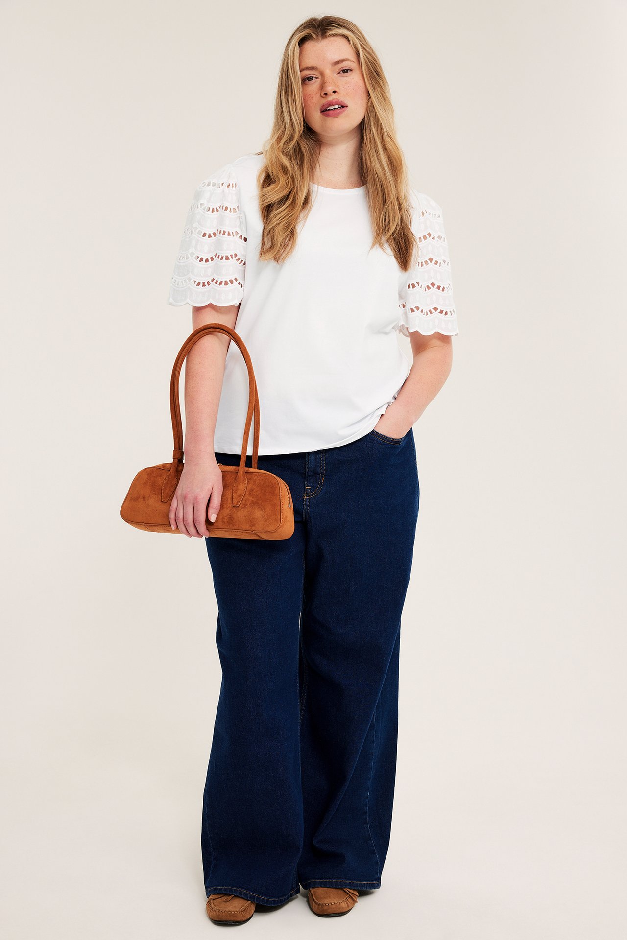 Edestäpäin: nainen valkoisessa broderie anglaise -topissa, tummansinisissä leveissä farkuissa, ruskeissa mokkasiineissa, pitää ruskeaa mokkalaukkua.