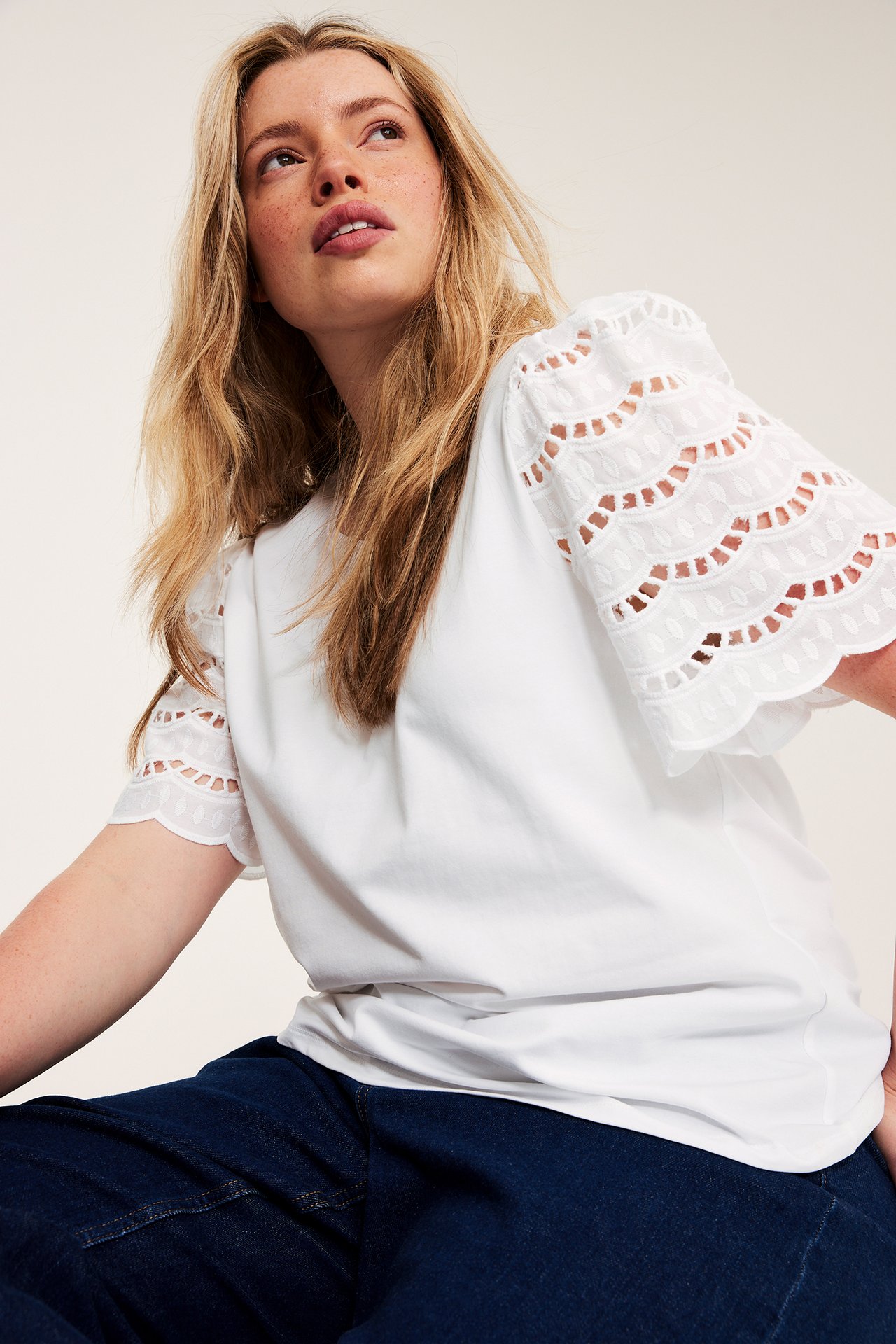 Naisen edestäpäin: valkoinen lyhythihainen toppi broderie anglaise -hihoilla ja tummansiniset farkut.