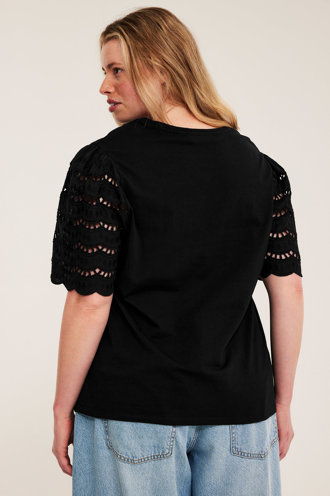 Naisen selkäkuva mustassa puuvillatopissa broderie anglaise -hihoilla ja vaaleansinisissä farkuissa.