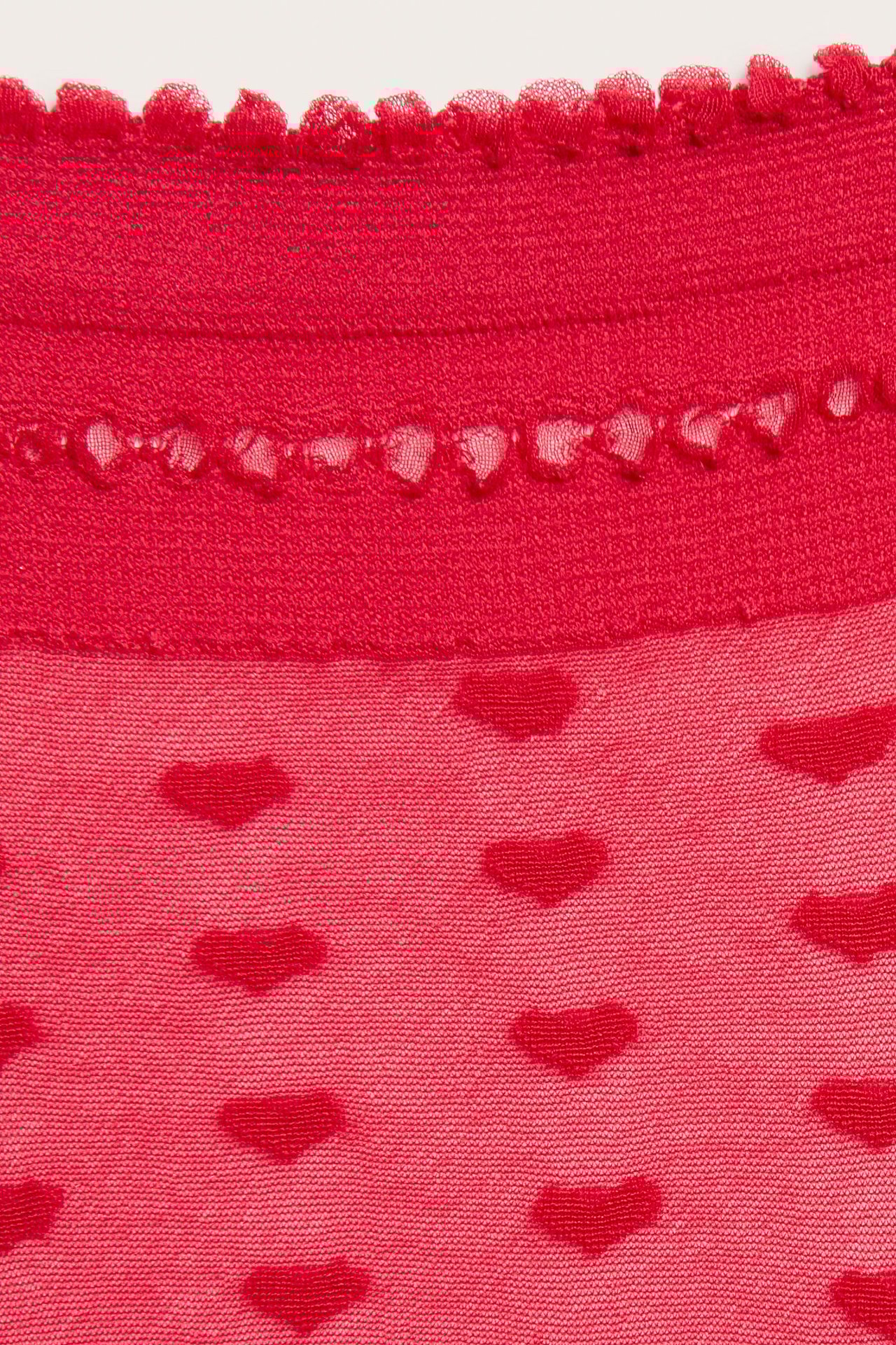 Nærbilde av en rød gjennomsiktig sokk med hjertemønster, bølget kant og perforerte detaljer.