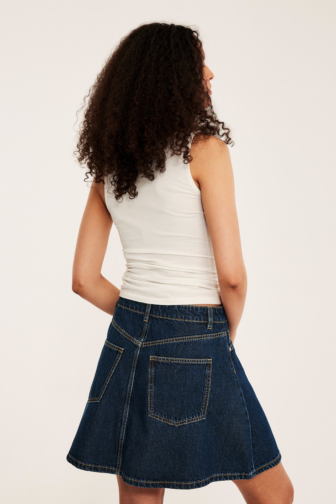 Ryggvy: kvinna i mörkblå A-linjeformad jeanskjol med gula sömmar och vitt linne.