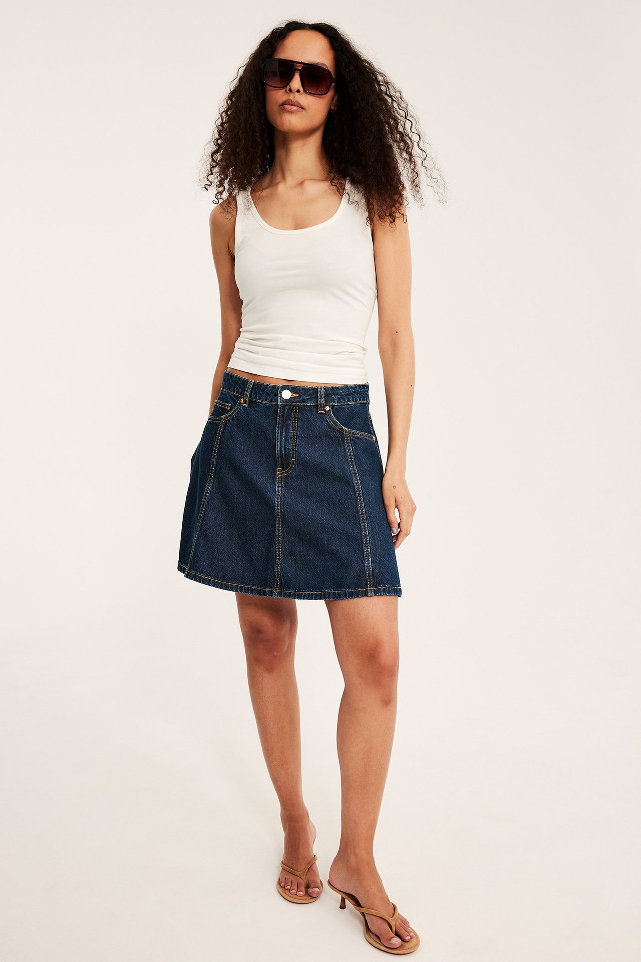 Framifrån, kvinna i mörk A-linjeformad jeanskjol, vit tanktopp, bruna solglasögon och sandaler.