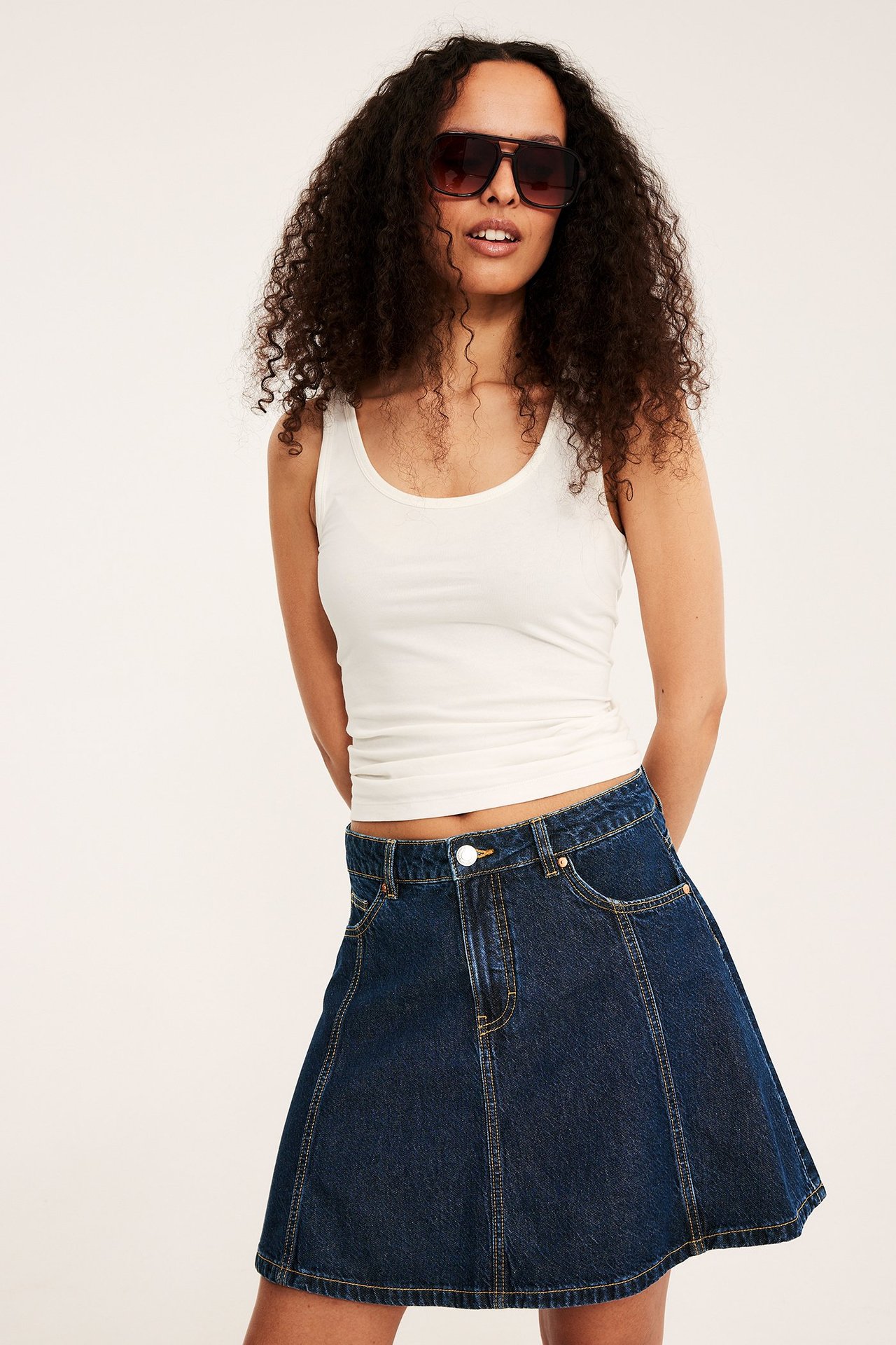 Frontvy: kvinna i mörkblå A-linjeformad jeanskjol, vitt linne och mörka solglasögon.