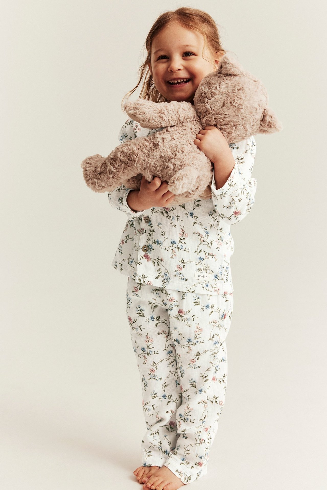 Frontvy: Smilende barn i offwhite blomstret pyjamas med knapper, holder en brun teddybjørn.