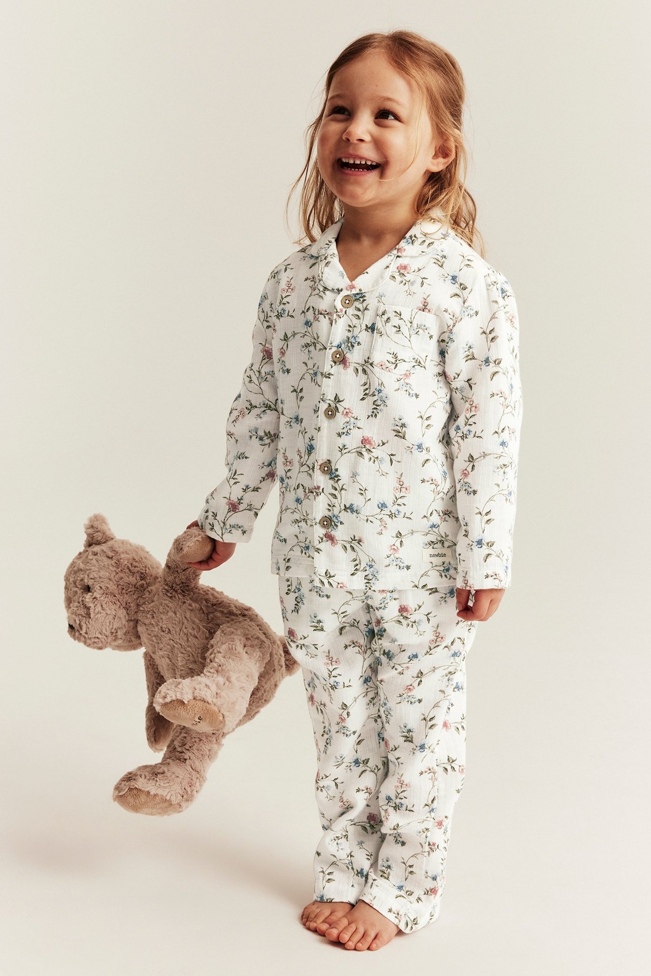 Edestäpäin, hymyilevä lapsi luonnonvalkoisessa kukallisessa kaksiosaisessa puuvillapyjamassa, pidellen ruskeaa nallekarhua.