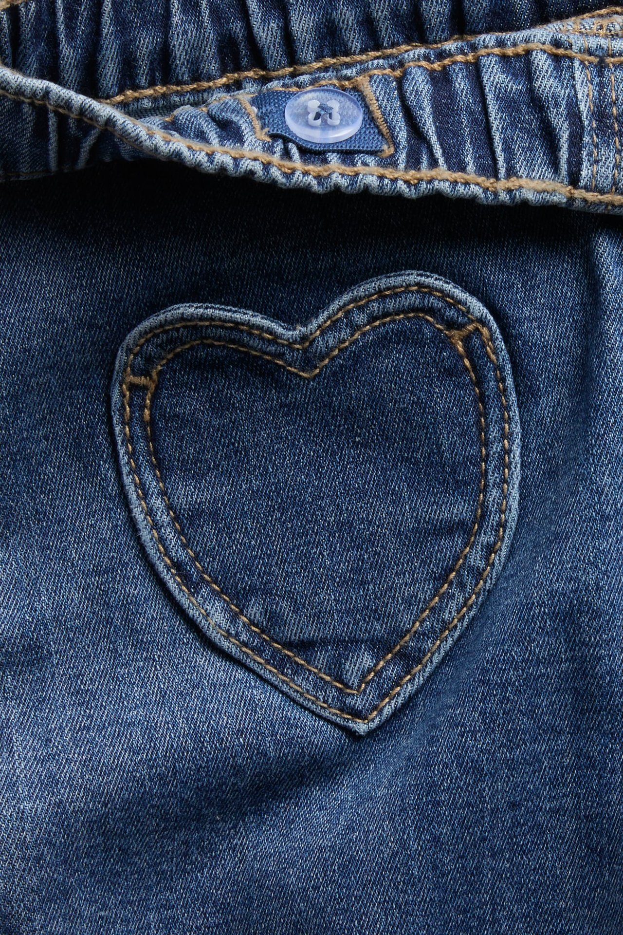 Närbild av blå denim med hjärtformad patch och en genomskinlig knapp på midjebandet.
