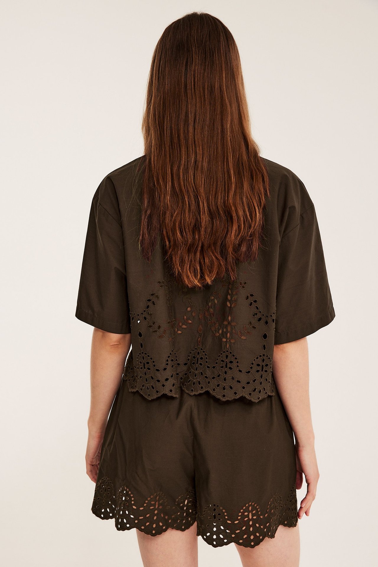 Ryggbilde av en kvinne i mørkebrune bomullsshorts og matchende skjorte, begge med broderie Anglais.
