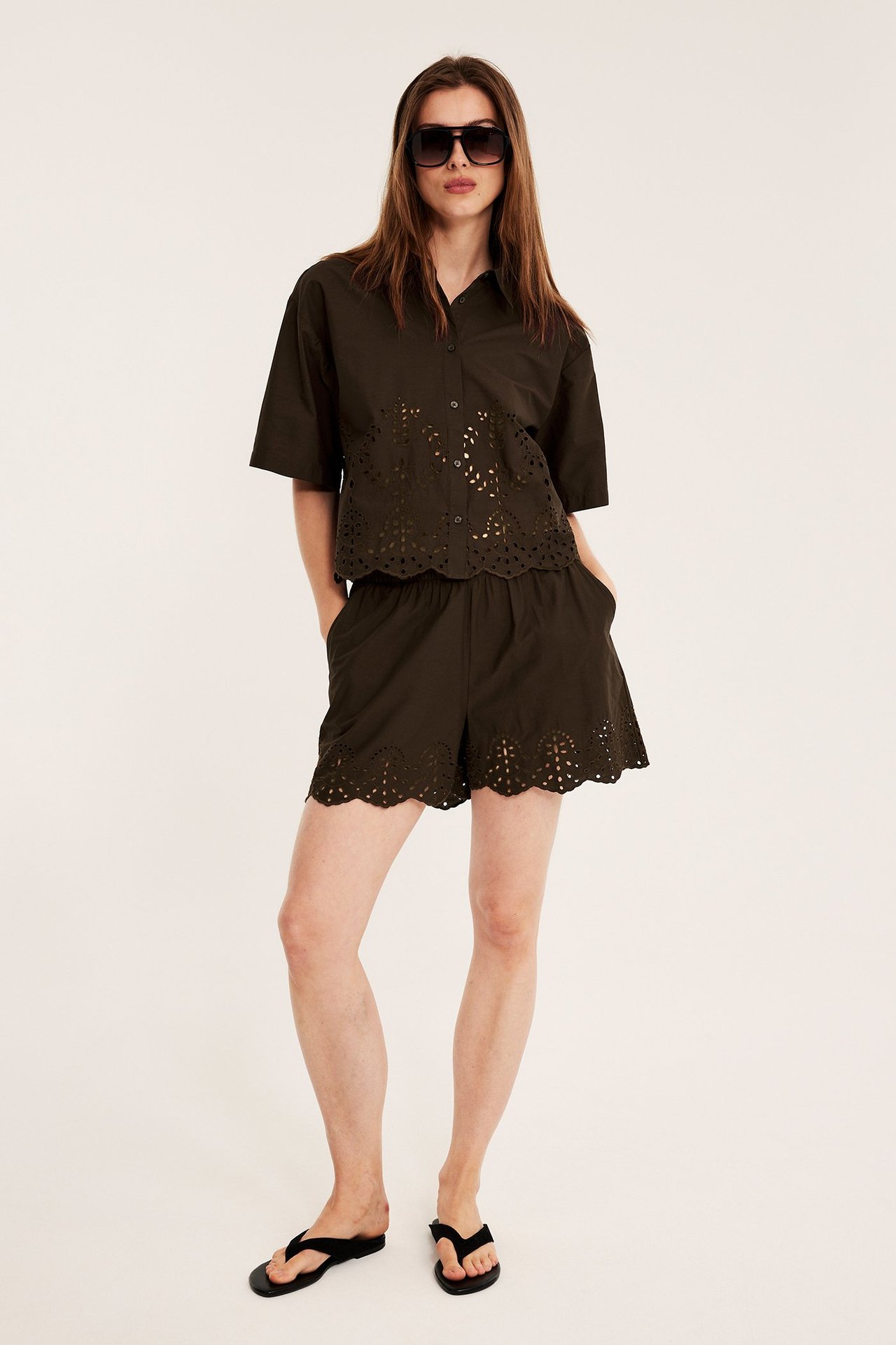 Edestäpäin: nainen yllään tummanruskeat puuvillapopliinishortsit englantilaisella kirjonnalla, yhteensopiva paita, varvassandaalit, aurinkolasit.