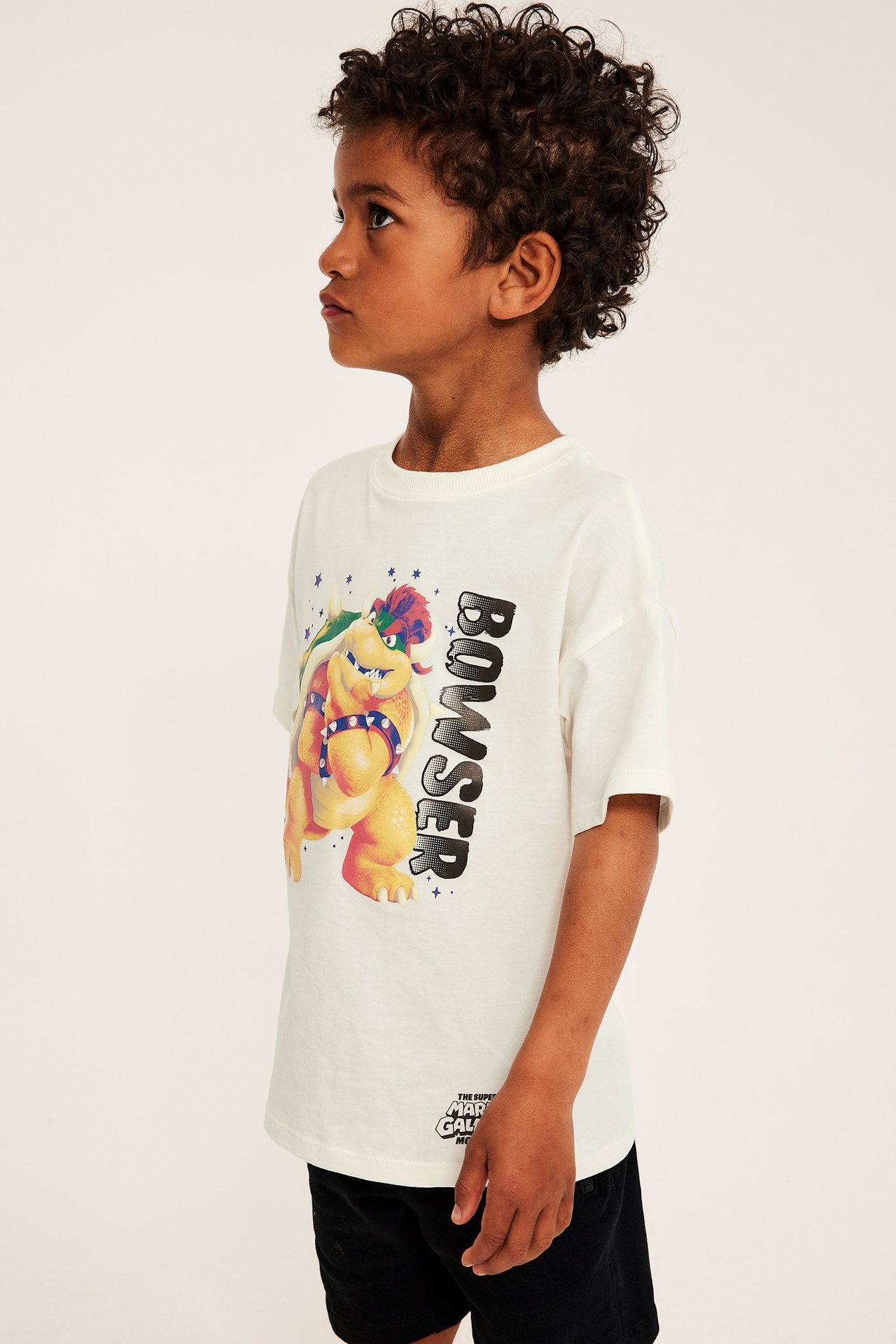 Widok z boku dziecka w kremowobiałym bawełnianym T-shircie z nadrukiem Bowsera, ze czarnymi szortami.
