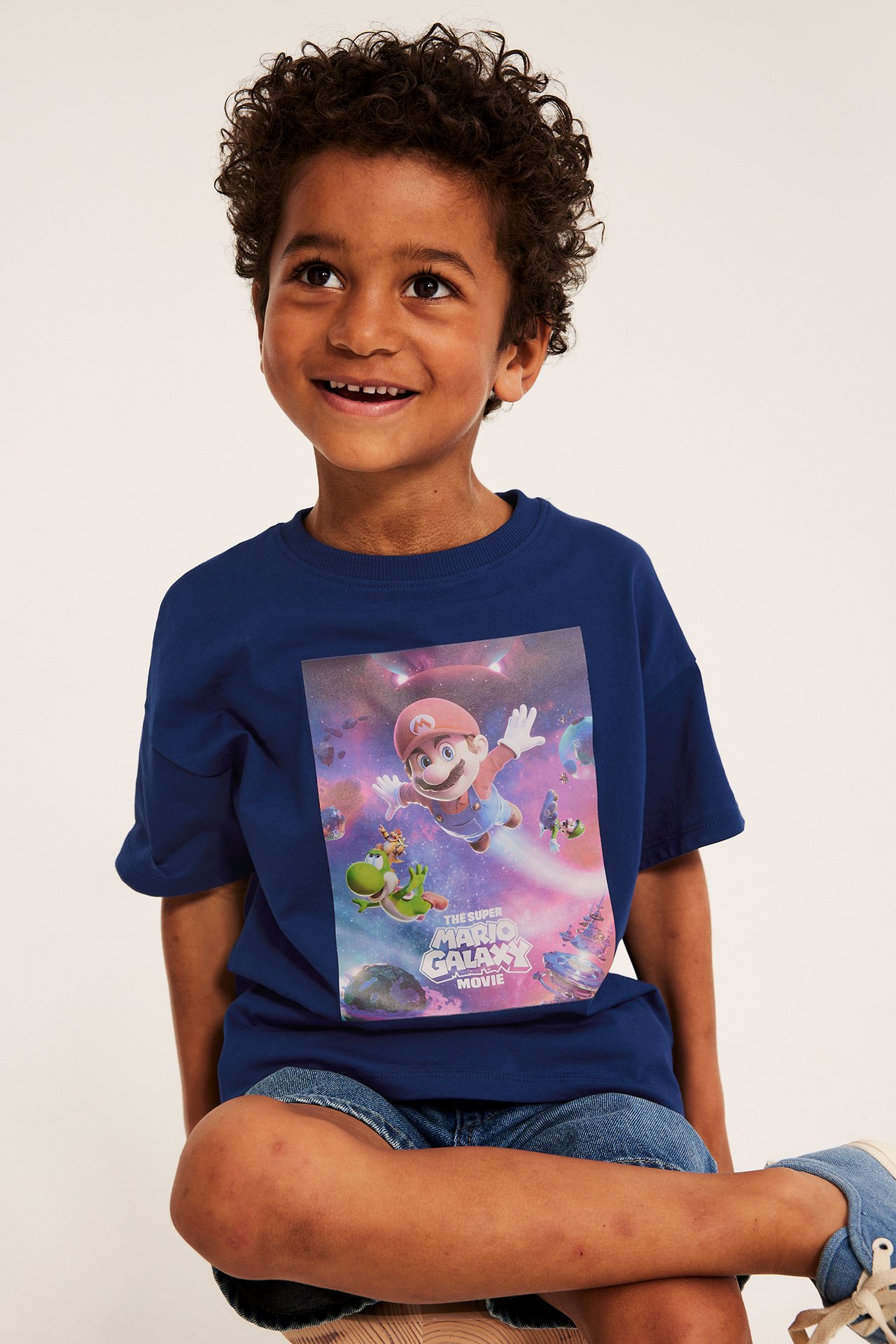 Widok z przodu dziecka w granatowym bawełnianym t-shircie z nadrukiem Super Mario Galaxy i szortach dżinsowych.