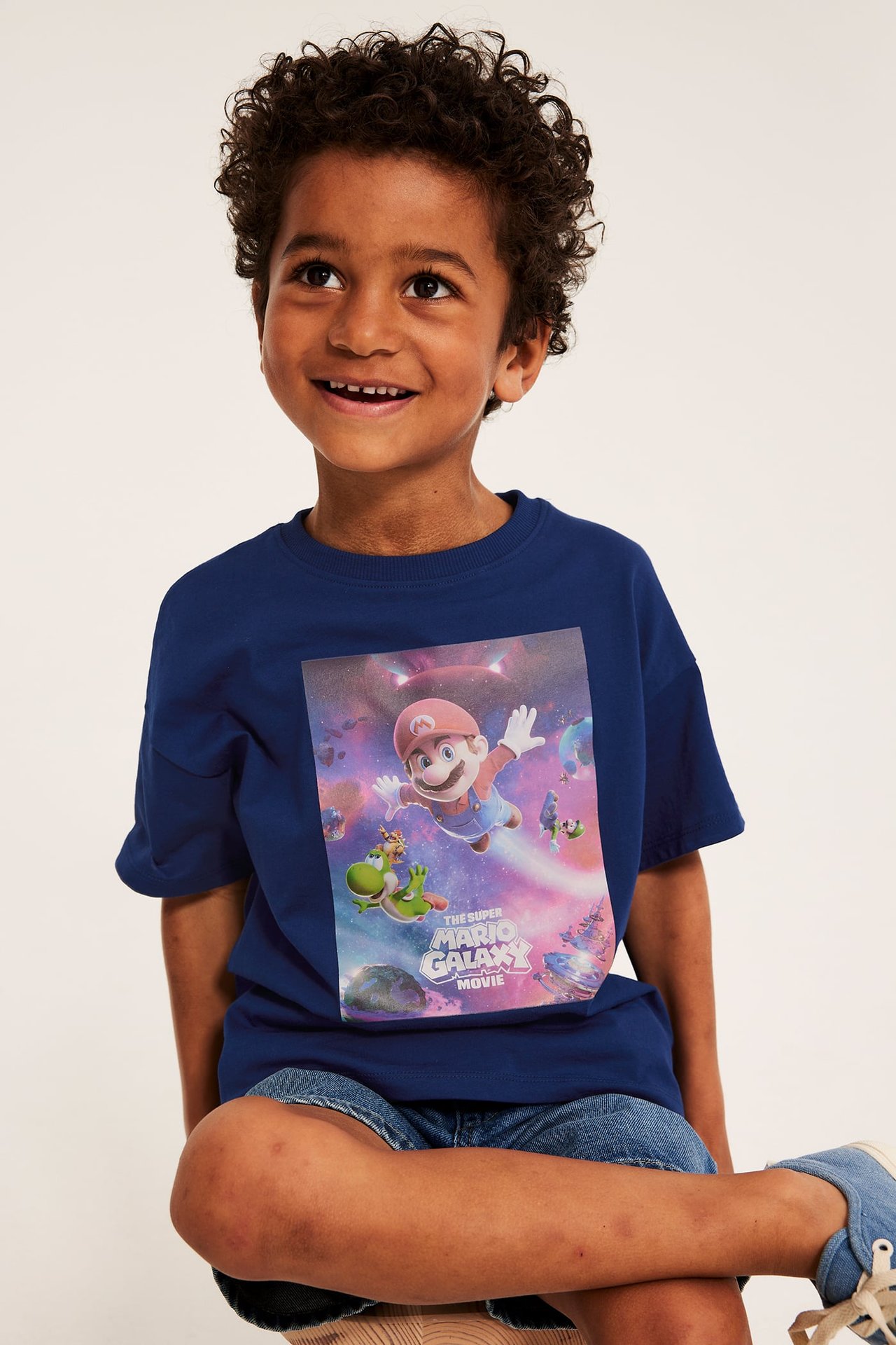 Frontvy av ett barn i en mörkblå Super Mario Galaxy bomulls-t-shirt med tryck och denimshorts.