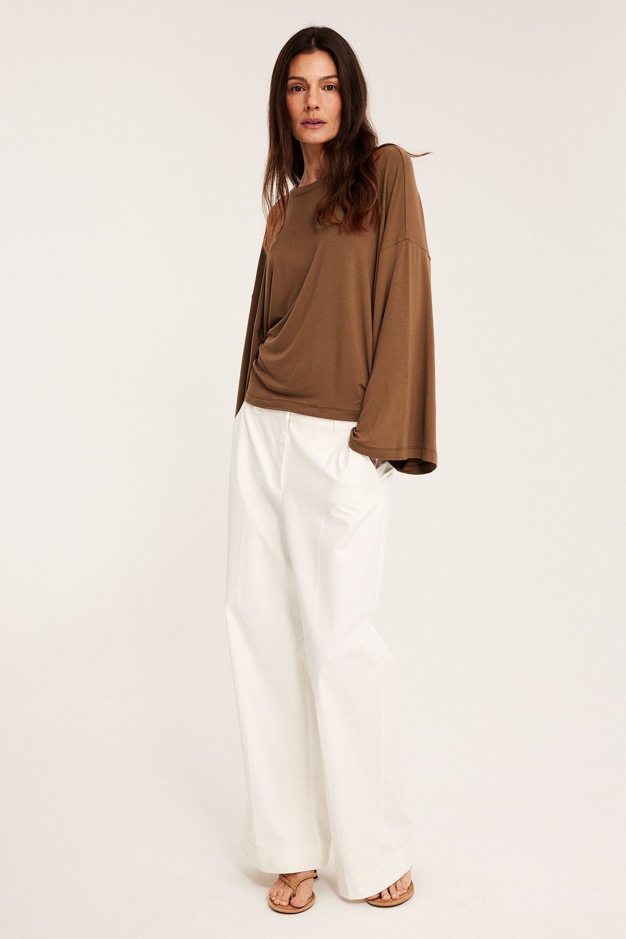 Kobieta w brązowym topie z szerokimi rękawami, białych szerokich spodniach i jasnobrązowych sandałach.
