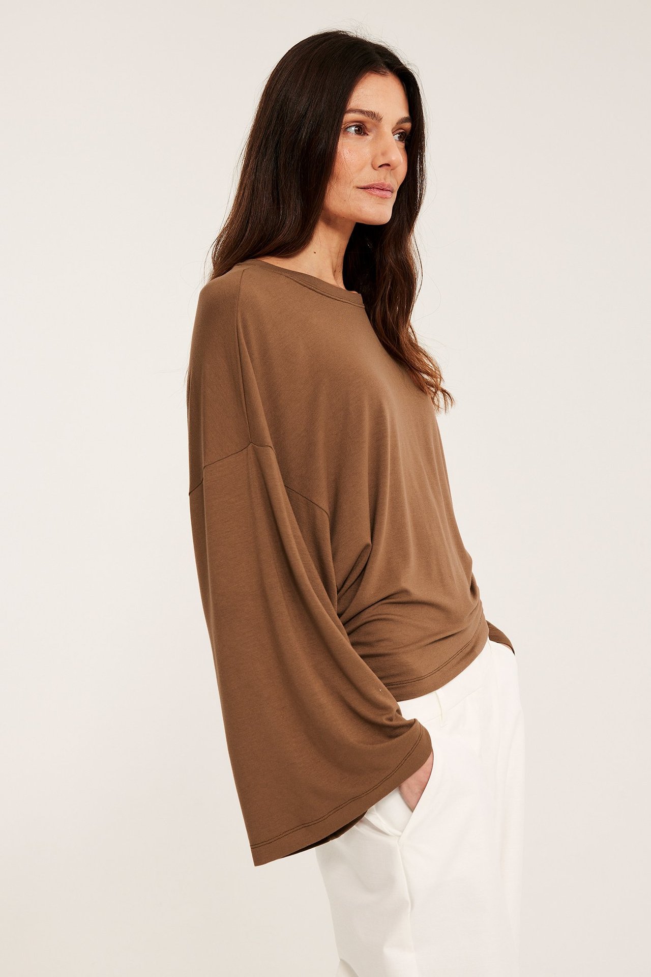 Sidovy av en kvinna i en brun draperad långärmad topp med vida ärmar och vita byxor.