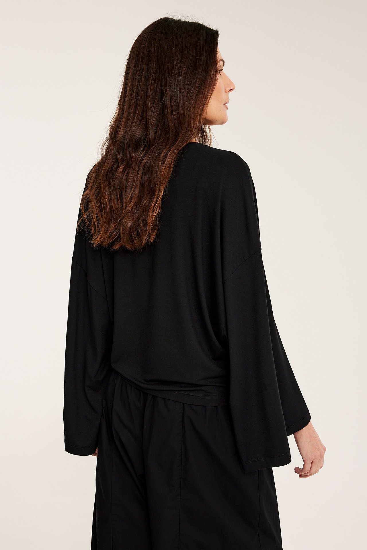Sidobakifrån: kvinna i svart draperad långärmad topp med vida ärmar och svarta byxor.